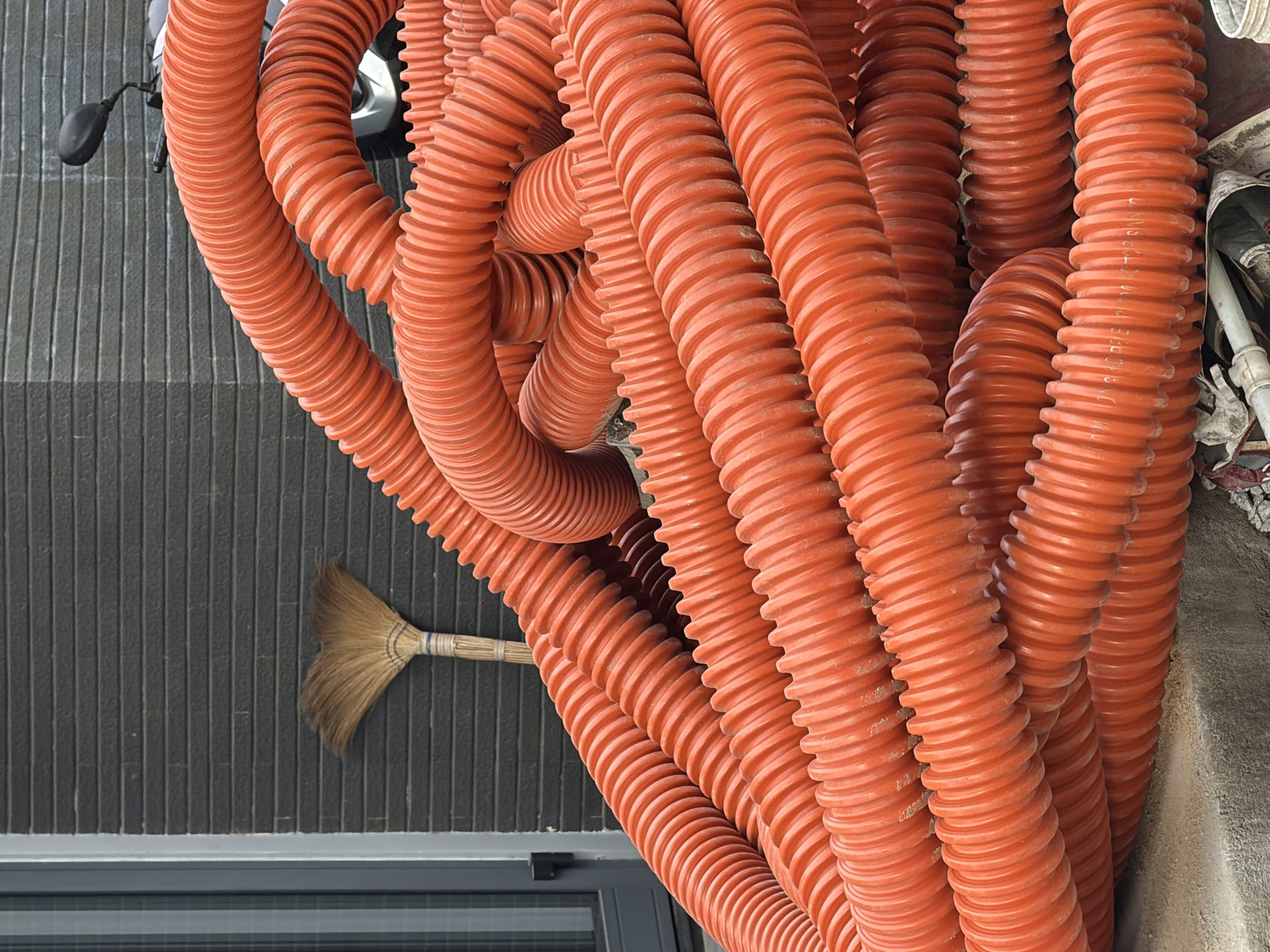 A high-angle shot depicts a dense cluster of orange corrugated plastic conduits or pipes. The pipes are arranged irregularly, filling the majority of the frame. Some pipes show signs of dust. To the left, a dark gray wall with vertical lines is partially visible, with the head of a small straw broom leaning against it. A sliver of a window is present at the bottom of the wall. The top of the image includes hints of other objects: an inflated bike tire and other construction debris. Given the presence of construction materials and elements in the background, the image appears to have been taken at a construction site in Đà Nẵng, Vietnam.