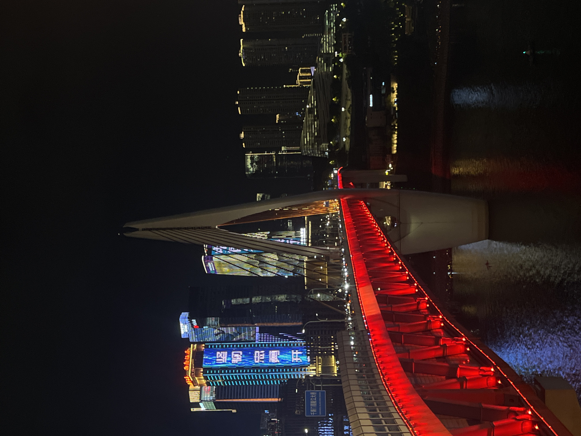The image captures a nighttime view of the Hongyadong Bridge in Yuzhong District, China. The bridge, illuminated with vibrant red lights along its railing and undercarriage, spans across a dark body of water. A prominent, white, pylon-like structure supporting the bridge rises from the center, with cables extending outwards. In the background, a cityscape of illuminated skyscrapers and buildings creates a dazzling urban panorama against the dark night sky. The water reflects the bridge's red glow and the lights from the city. There are no visible people or specific actions depicted in the scene.