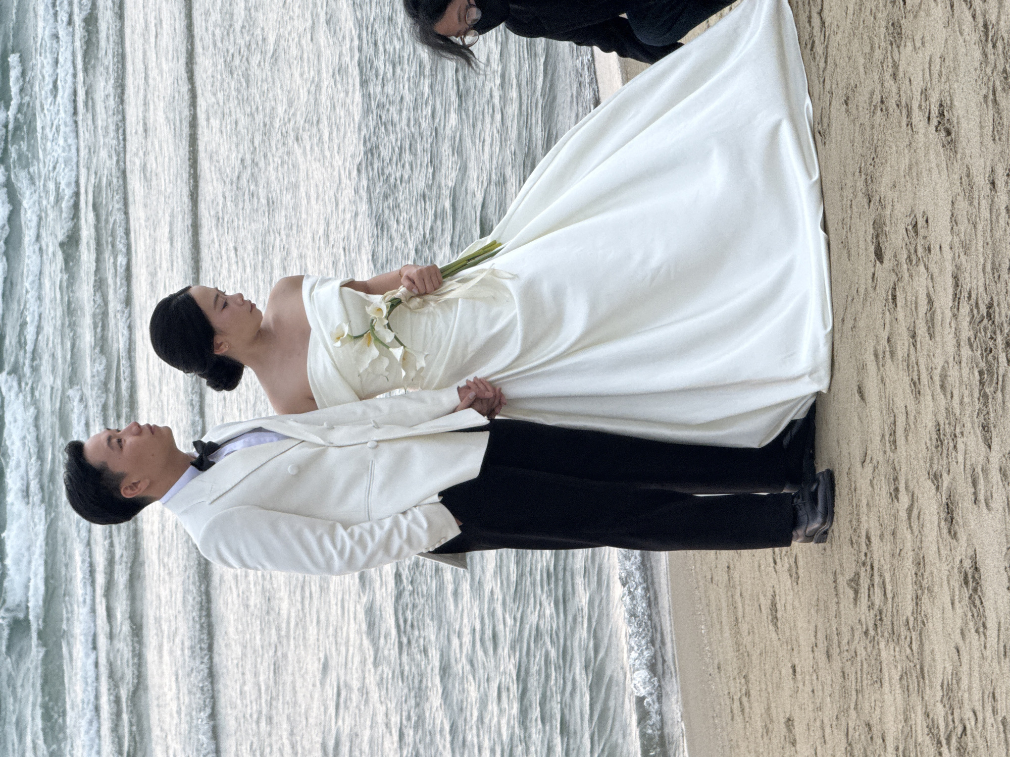 A wedding photo captured at a beach in Đà Nẵng, Vietnam shows a couple in formal attire. The bride wears a white, strapless wedding gown and holds a bouquet of white calla lilies. The groom is dressed in a white tuxedo jacket, black pants, and a black bow tie. The couple stands near the shoreline with the ocean waves visible in the background. A person wearing glasses in black clothing stands nearby. The ground is sandy.