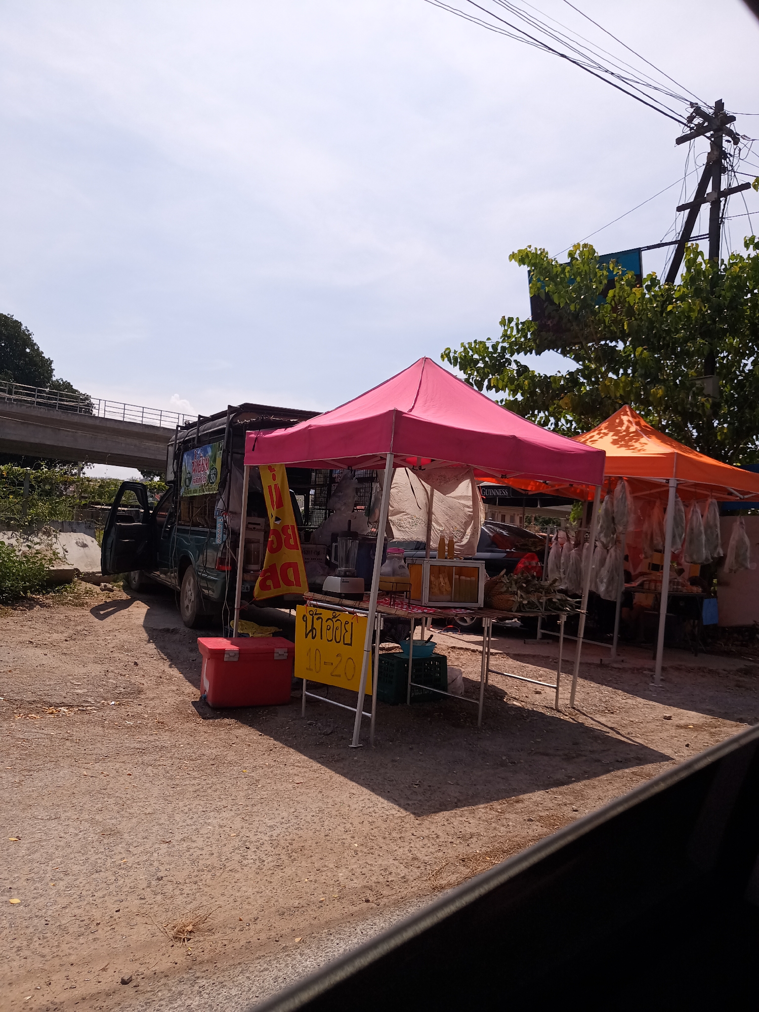 The image shows a roadside scene in Hua Hin City Municipality, Thailand. A dark green pickup truck, modified to serve as a mobile vendor stall, is parked on a dirt patch next to a paved road. The driver-side door is open. Two pop-up tents, one pink and one orange, are erected next to the truck, indicating a marketplace setup. The tents appear to house food items. A red cooler sits on the ground near the truck. A yellow sign with Thai script and the numbers "10-20" is visible near the pink tent.
In the background, a concrete overpass is visible, and a utility pole stands with multiple wires. The sky is bright with scattered clouds, suggesting daylight conditions. There is no evidence of pedestrian or vehicular traffic nearby.