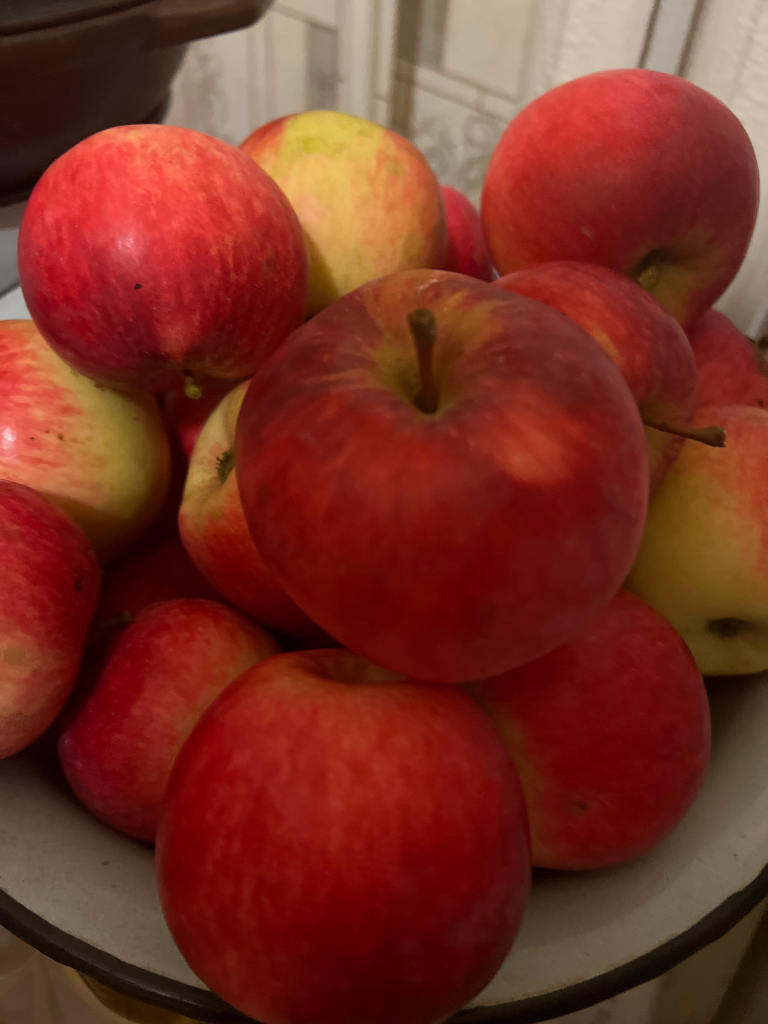 A close-up, overhead shot shows a bowl filled with red and yellow apples. The apples are clustered together, with some partially obscured by others. The lighting is dim, casting shadows on the fruit. The background is out of focus, but appears to be a wall with a textured pattern.
