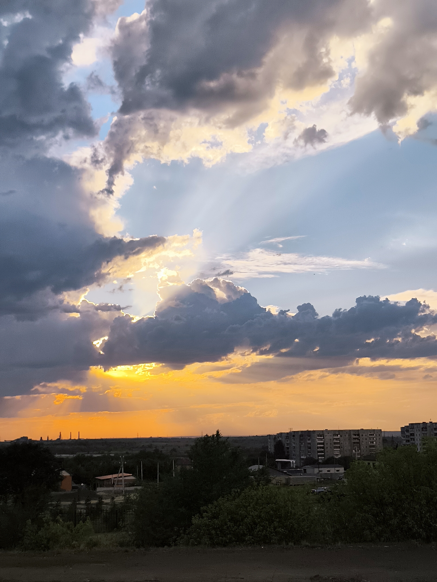 A wide-angle, slightly low-angle view captures a dramatic sunset over an urban landscape. The sky is dominated by heavy, dark gray clouds interspersed with bright, sunlit patches that cast dramatic crepuscular rays downwards. The lower portion of the sky is a vibrant orange and yellow, indicating the sun is low on the horizon. In the distance, silhouetted against the bright sky, are industrial structures, possibly a power plant with several tall stacks. Closer to the foreground, a cluster of multi-story apartment buildings, characteristic of Soviet-era architecture, are visible. Dense green foliage in the mid-ground obscures some of the lower parts of the buildings and the landscape, while the immediate foreground consists of a dark, unpaved surface. The scene suggests an industrial city in Kazakhstan, likely within the Qarağandı City Administration area, experiencing the transition from day to night.