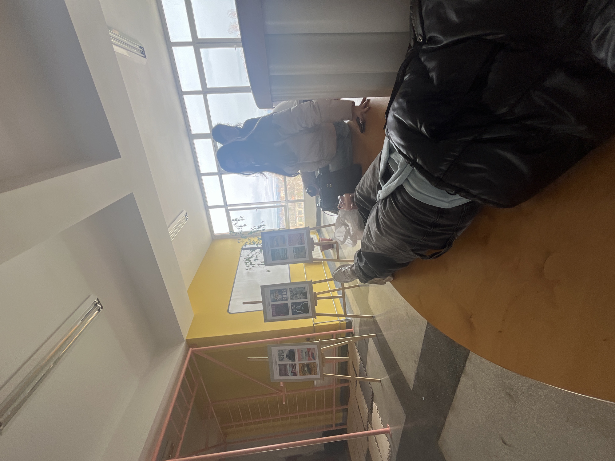 People are in a well-lit interior space with a view out to a city, possibly Kyustendil, Bulgaria. Artwork is displayed on easels. Some of the people are interacting with the artwork, and the environment includes pink scaffolding.