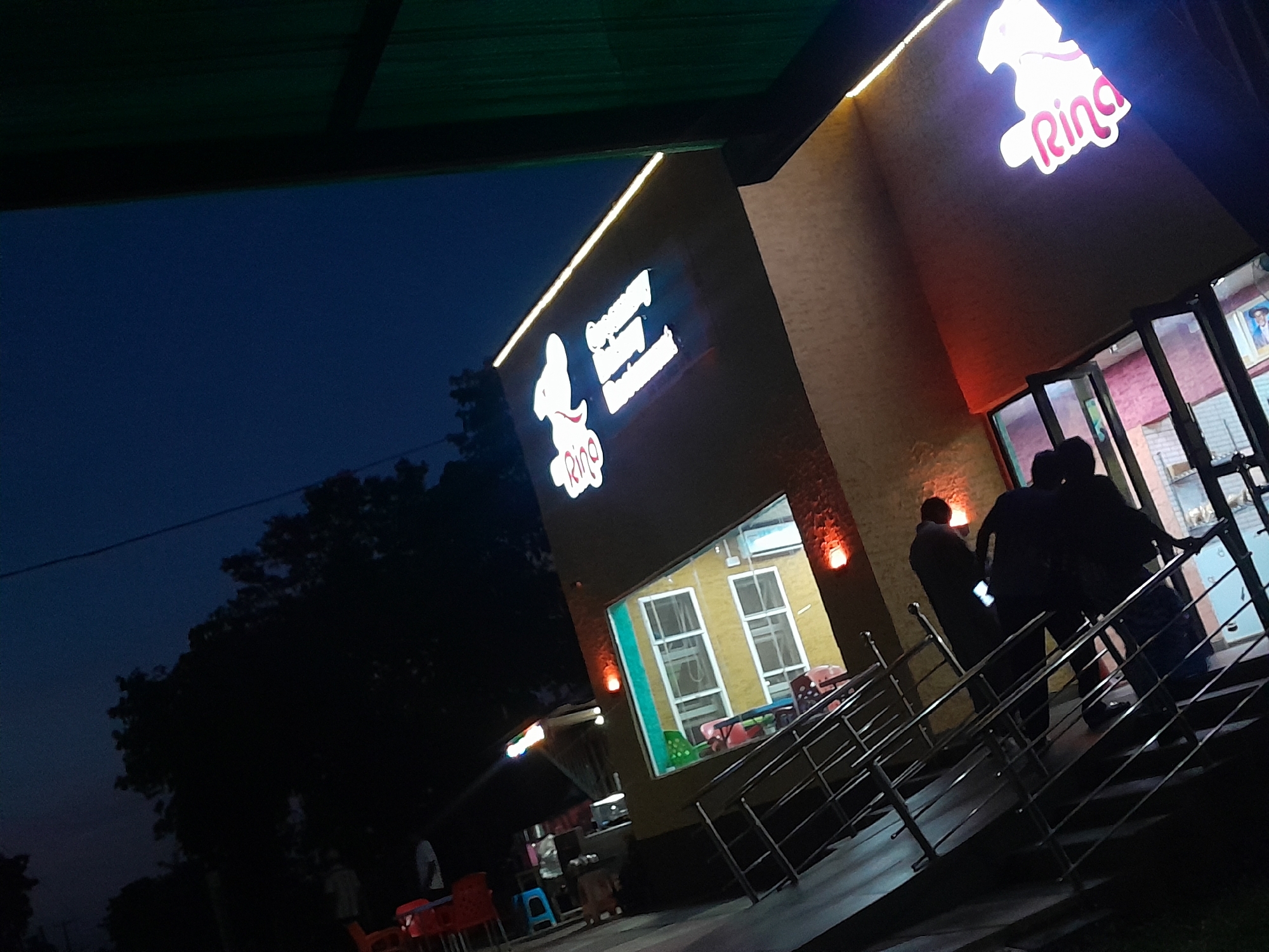 This media file captures an exterior view of a brightly lit restaurant named "Rino" at dusk or night in Ilorin, Nigeria. The building features a yellowish-tan facade with prominent strip lighting along its roofline and warm orange wall sconces.
The main subject is the restaurant building, displaying a large, illuminated sign with a distinctive white logo that resembles a stylized animal or chicken. Below this logo, the word "Rino" is written in a playful pink font. Above the "Rino" logo, there is text indicating menu items, which appear to be "Crispy Chicken" and "Burgers & Sandwiches." Another smaller "Rino" text is also visible.
In the foreground, a modern metallic railing system leads up to the restaurant's entrance, where several people are gathered. Their figures are largely silhouetted against the interior and exterior lighting, making their specific actions unclear, though they appear to be either entering or exiting. One person on the steps is looking at a mobile phone.
Through large windows on the building, elements of the interior dining area, including tables and chairs, can be seen. On the left side of the image, additional outdoor seating with red and blue chairs and tables is visible on what looks like a patio or sidewalk area. The background features a dark blue sky, indicative of evening, with the silhouettes of large trees. A dark green overhang is present in the upper left corner of the frame. The scene depicts a lively commercial establishment at night.