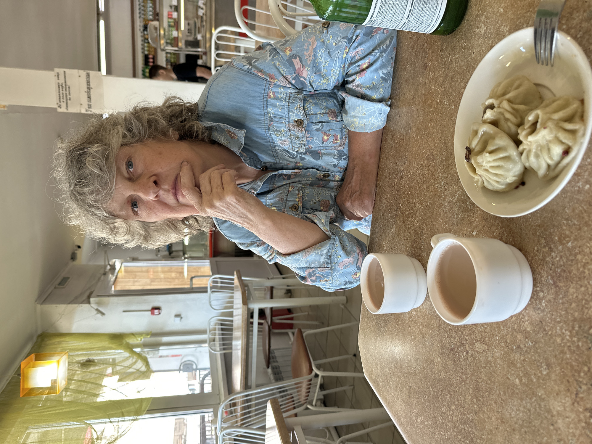 An elderly Caucasian woman with curly gray hair is seated at a table in a dining establishment, possibly a canteen or cafe. She is wearing a blue denim shirt with a floral pattern. Her right hand is resting on her chin, and she is looking directly at the camera with a neutral expression. On the table in front of her are two white mugs filled with a light brown beverage, a small white plate containing three dumplings, and a green bottle. The background shows tables and chairs, windows with yellow curtains, and a serving counter with bottles and food items. The general atmosphere suggests a casual dining setting in Gorno-Altaysk, Russia.