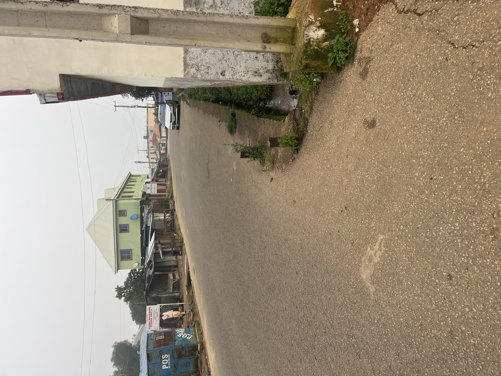 The image depicts a street scene in Jos, Nigeria, characterized by a paved road with a slight incline. On the left side of the road, there are various small businesses housed in structures that appear to be made of corrugated metal and wood. One prominent blue structure has "POS PAY YOUR NEPA BILL" written on it, indicating a point-of-sale service for utility payments. A person in a light-colored traditional outfit stands near these businesses. Further down the street, a two-story building painted light green with a white roof is elevated on stilts, featuring multiple windows.
On the right side of the road, a concrete drainage channel runs alongside the pavement, with overgrown vegetation in and around it. A weathered concrete pillar, possibly part of a building or a utility pole, stands prominently in the foreground on the right, showing signs of moss and age. A silver sedan is parked on the right side of the road, facing away from the viewer, with signage visible on the building behind it. The sky is overcast, suggesting a hazy or possibly rainy atmosphere, and the overall environment appears to be a busy commercial and residential area in Jos.