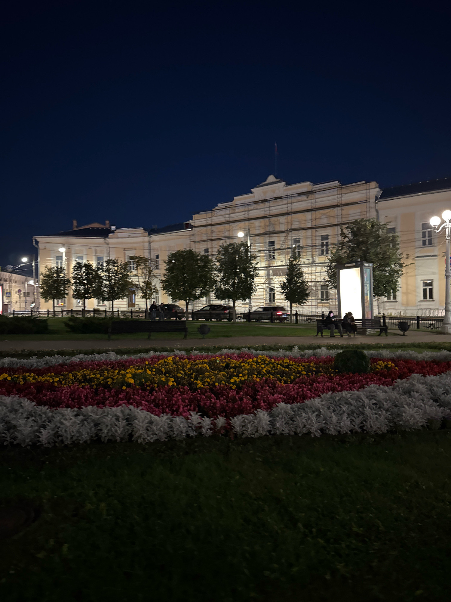 This image depicts a nighttime scene in Tver, Russia, focusing on a building with scaffolding, a flowerbed, and a park area. The building, likely a historic structure due to its architecture and the scaffolding suggesting ongoing renovation, is illuminated by artificial light, revealing its pale yellow facade with multiple windows. A flag flies from the roof.
In the foreground, a meticulously arranged flowerbed creates a vibrant display of colors: white, pink, yellow, and red flowers are planted in distinct rows, forming a striking visual contrast against the dark green grass and the dim evening sky. A few trees are strategically placed along the perimeter of the park.
Beyond the flowerbed, a paved walkway is visible, bordered by a metal fence. Several park benches are present, with two individuals seated on one of them, seemingly engaged in conversation or observation. A tall, illuminated advertising stand stands near the benches. Several cars are parked or driving along a road behind the fence, their headlights and taillights subtly visible. Streetlights are also present, casting pools of light on the walkway and the building. The overall atmosphere is tranquil, characteristic of a city park at dusk or early evening.