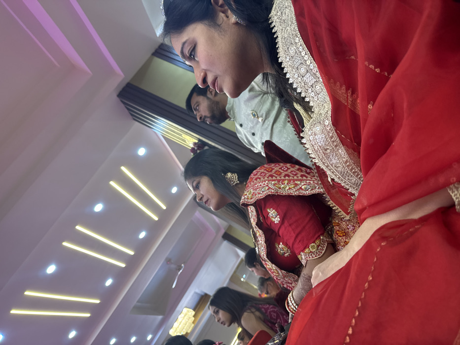 A group of people are dressed up in traditional Indian clothing, seemingly observing or participating in a ceremony. The woman in the foreground wears a red outfit with intricate gold embroidery and a delicate silver tiara. Behind her, another woman is dressed in a similar red ensemble. A man is visible with a pale green shirt. The ceiling is modern, with linear lighting fixtures. The photo appears to be taken from a low angle, capturing the scene from below.