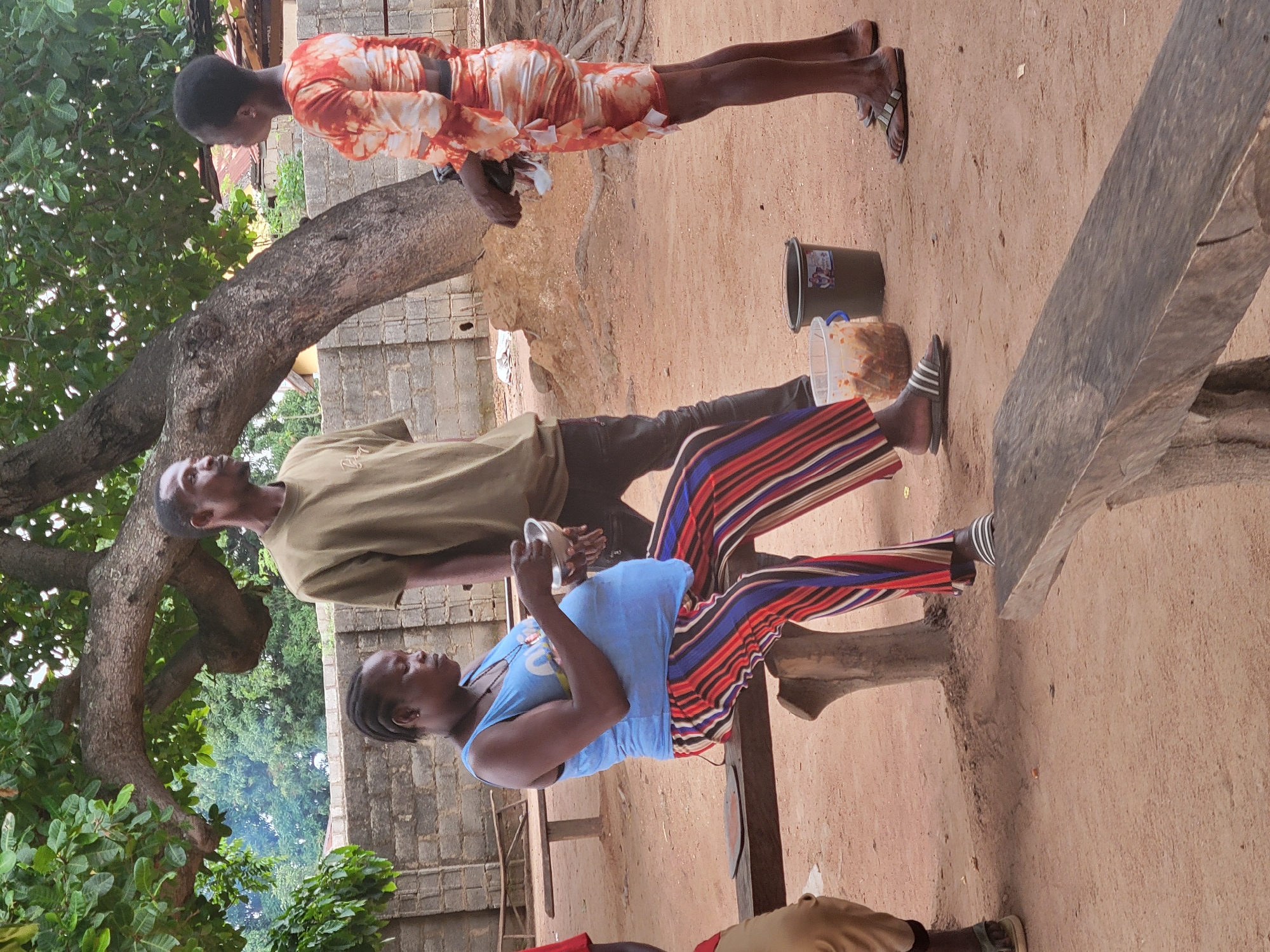 This outdoor scene in Madalla, Nigeria, captures several people under the shade of a large, gnarled tree with green foliage. The ground is dusty and sandy, suggesting a dry environment.
Three adults are prominently featured. A woman wearing an orange and white patterned short dress stands on the right, looking downwards. A man in a light khaki or olive green t-shirt and dark trousers stands to her left, gazing upwards or into the distance. Another woman, wearing a blue sleeveless top and striking striped trousers (red, blue, white, and black stripes), is seated on a low wooden bench-like structure, holding a small white bowl. Her gaze is directed towards the ground or the items beside her. Partially visible at the very bottom, another person wearing a red shirt is seated.
On the ground near the seated woman, there's a dark-colored bucket with a faded label, and a clear plastic container filled with what appears to be fried snacks or nuts. All individuals are wearing sandals or flip-flops. In the background, a building constructed of cinder blocks is visible. The bright lighting suggests it is daytime, likely midday or afternoon, with clear skies. The overall mood appears casual and relaxed, with individuals seemingly interacting or resting. No decipherable text is clearly visible on any objects.