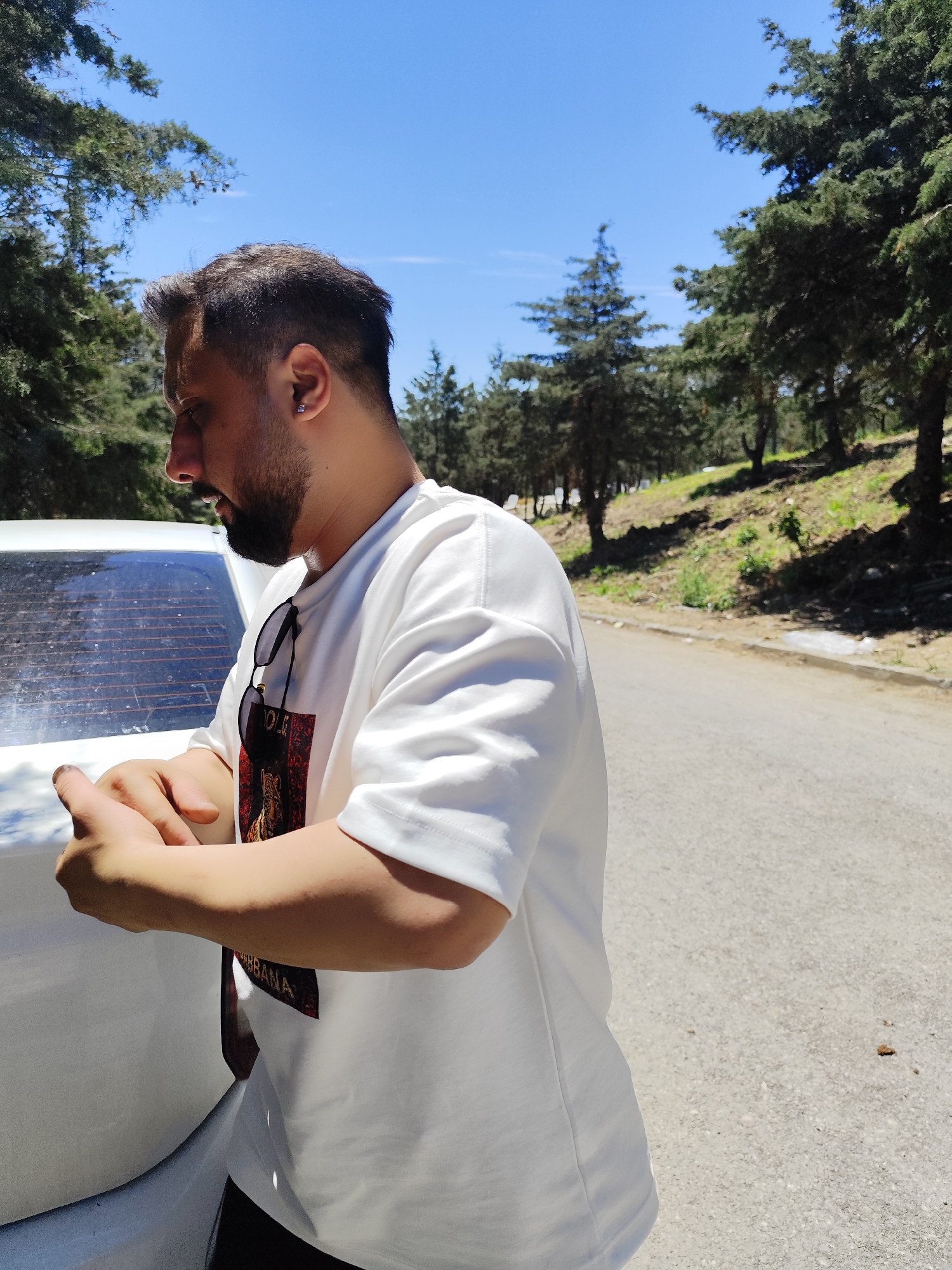 A medium shot shows a man in profile standing beside the back of a white car on a sunny day. The man has dark hair, a beard, and wears a white t-shirt with a graphic print on the front, and sunglasses hang from the shirt. He's touching his hands together, possibly looking at his nails. The car is parked on the side of a road. Behind the car and man, there are several tall evergreen trees against a bright blue sky. The road appears to be in a park or wooded area. Based on the terrain and tree types, the location is potentially within Sarıyer, Turkey, a district known for its natural beauty and forested areas.