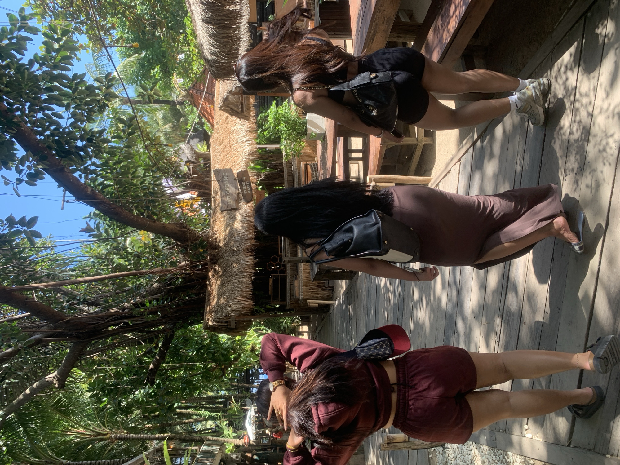 Three women walk along a wooden boardwalk lined with tropical foliage and thatched-roof structures in the city of North Kuta, Indonesia. The woman in the foreground on the left wears a maroon hoodie and shorts, with her hands on her head. The woman in the middle wears a long, brown dress and carries a black backpack. The woman on the right wears black shorts and a black top, also carrying a black bag. Sunlight filters through the trees, casting dappled shadows on the boardwalk. The environment suggests a tropical resort or market setting.