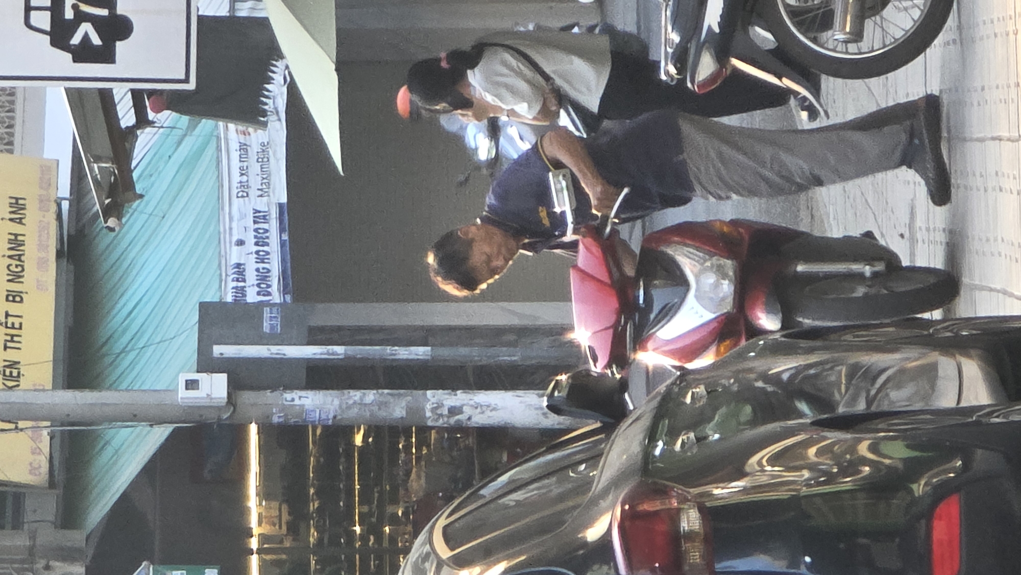 The image shows a street scene, seemingly in Nha Trang, Vietnam, as indicated by the signage in Vietnamese language. A man is positioned near a red and silver scooter, his stance suggesting he might be about to mount or dismount. Another person, possibly a woman, stands behind him, looking down. A parked black car partially obscures the lower part of the scooter. In the background, there is a green awning and various signs, including one advertising Maxim Bike. A street light pole is in the middle of the frame.