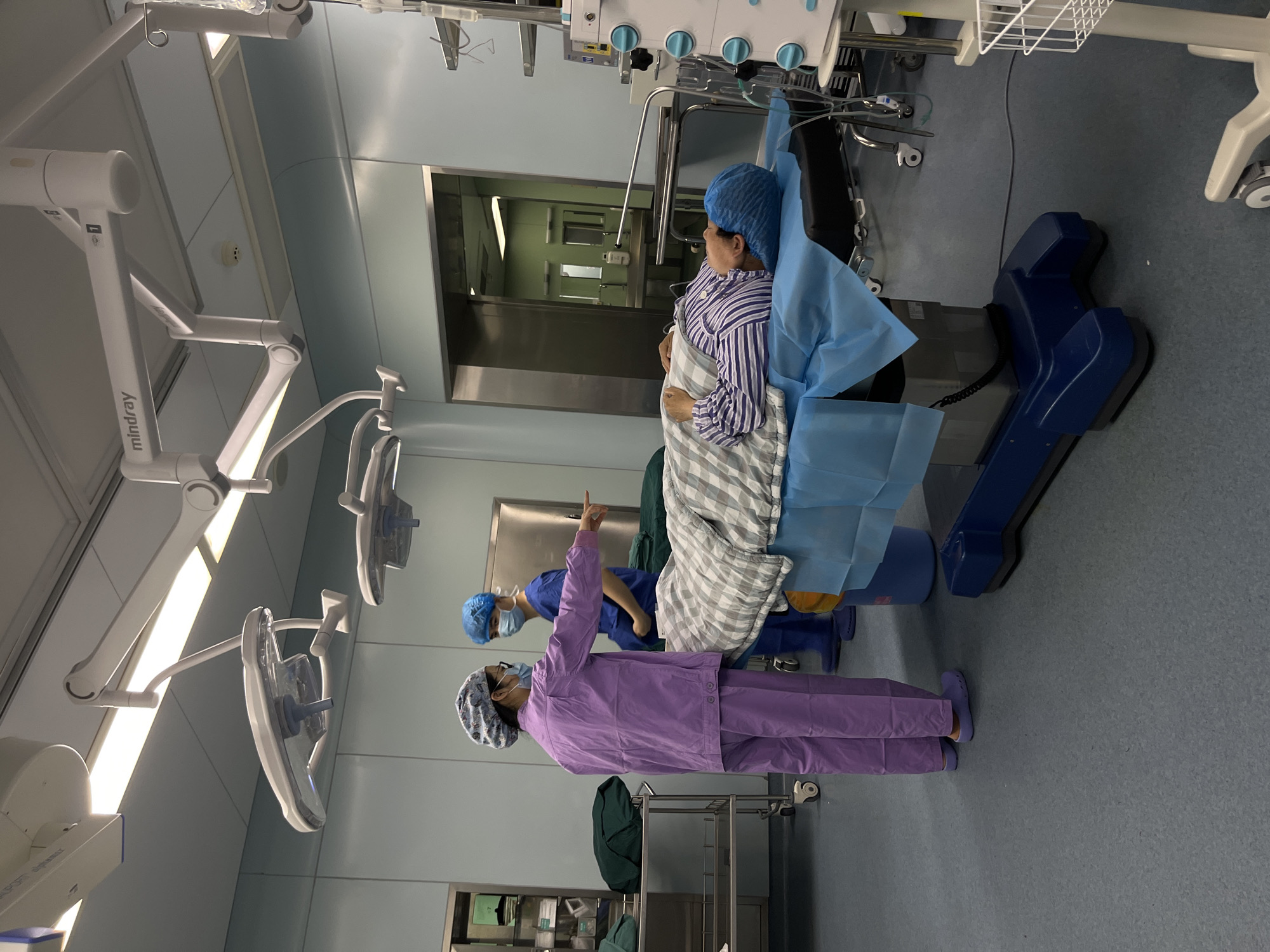 The image shows a surgical operating room. A patient lies on an adjustable examination table, covered with a blue surgical drape and a gray blanket with white stripes. Two healthcare workers are present. One, wearing lavender scrubs, stands pointing upward, with a surgical cap and glasses. The second healthcare worker wears blue scrubs and a surgical cap, also wearing a face mask. Medical equipment is visible, including surgical lights labeled "mindray," an equipment tower with controls and a wire basket, and a stainless-steel cabinet. The floor is gray. A metal door frame is visible. There is no specific evidence to confirm that the photo was taken in Yuzhong District, China.