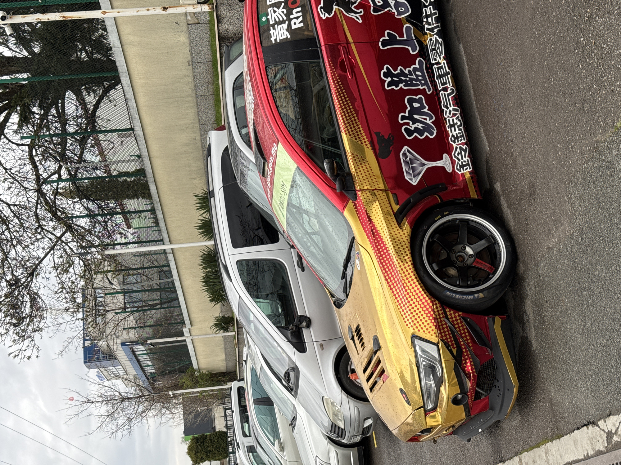 The image depicts a row of parked cars on a street, likely in a commercial or residential area. The most prominent car is decorated with a unique red and gold livery, featuring diamond graphics and Asian lettering. The other vehicles are standard silver and white models, with some parked closely together. The scene is in Sofia, Bulgaria, based on identifying features like the Asian lettering on the car that contains characters indicating a business or organization linked to Macau, a special administrative region of China, and given there is an online presence for a car club with similar vehicles in Sofia, Bulgaria. The weather appears overcast, and there is a fence, bare trees, and a building visible in the background. The environment seems urban, and the overall impression is of a typical day on a city street.