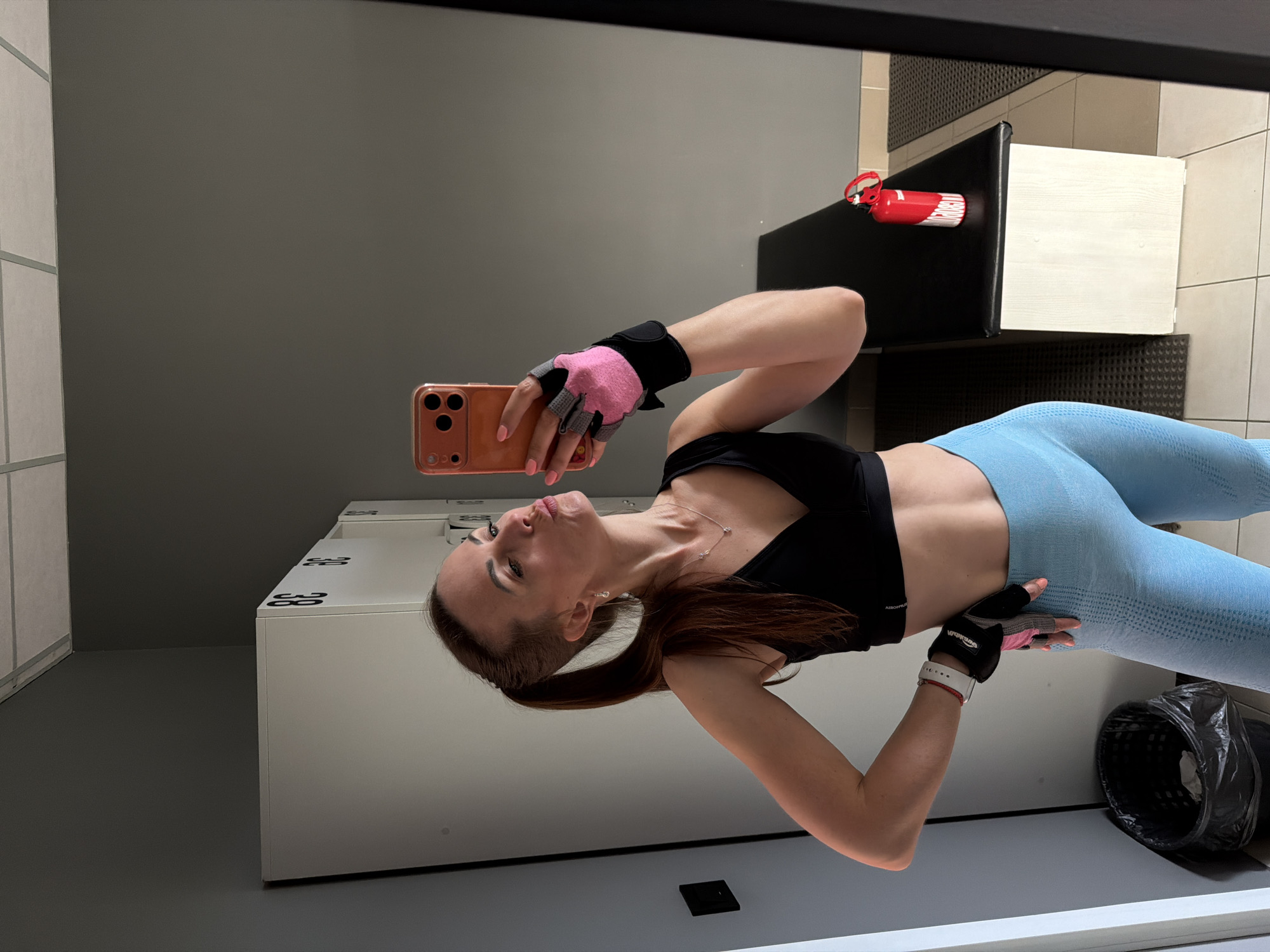 A woman with brown hair in a ponytail, wearing a black sports bra and light blue leggings, takes a selfie in a gym locker room. She is wearing pink and gray workout gloves and has her right hand on her hip. A smartphone is visible in her right hand, and she is puckering her lips towards the camera. In the background, there are white lockers with black numbers, a black trash can, and a black bench with a red water bottle. The floor is tiled, and the walls are gray. The scene appears to be in a public locker room.