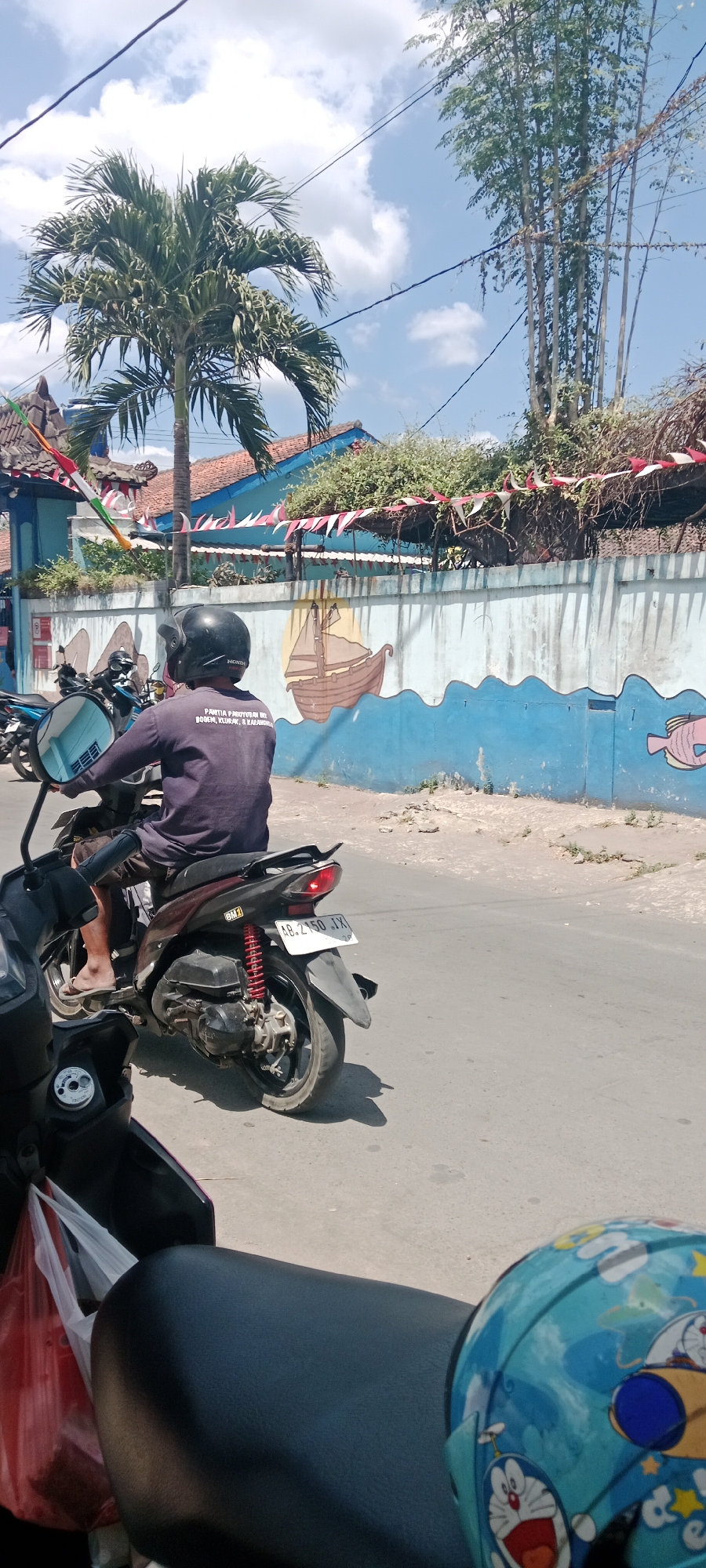 In the city of Purwomartani, Indonesia, on a bright, sunny day, a person wearing a dark long-sleeved shirt, shorts, and a black helmet rides a maroon scooter. The scooter has a license plate that reads "AB 2150 JX". The person is on a paved road, and to their right is a low wall with a mural. The mural depicts a blue ocean with white waves, a brown sailboat with a yellow sail, and a pink fish. Behind the wall, there are trees, including a palm tree to the left and tall, thin trees to the right, with buildings and red and white triangular flags decorating the top of the wall. In the foreground, the seat of another scooter is visible, along with a blue helmet adorned with cartoon characters.