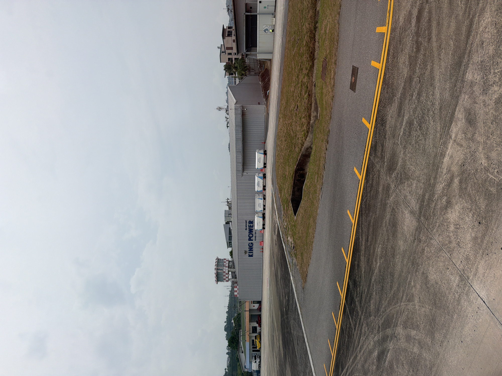 The image shows a view of the King Power building at Phuket International Airport in Mai Khao, Thailand. A portion of the runway is visible to the right of the building with yellow lane markings. The sky is overcast. Several service vehicles are parked alongside the building. The surrounding area appears to be a mix of industrial and residential properties.