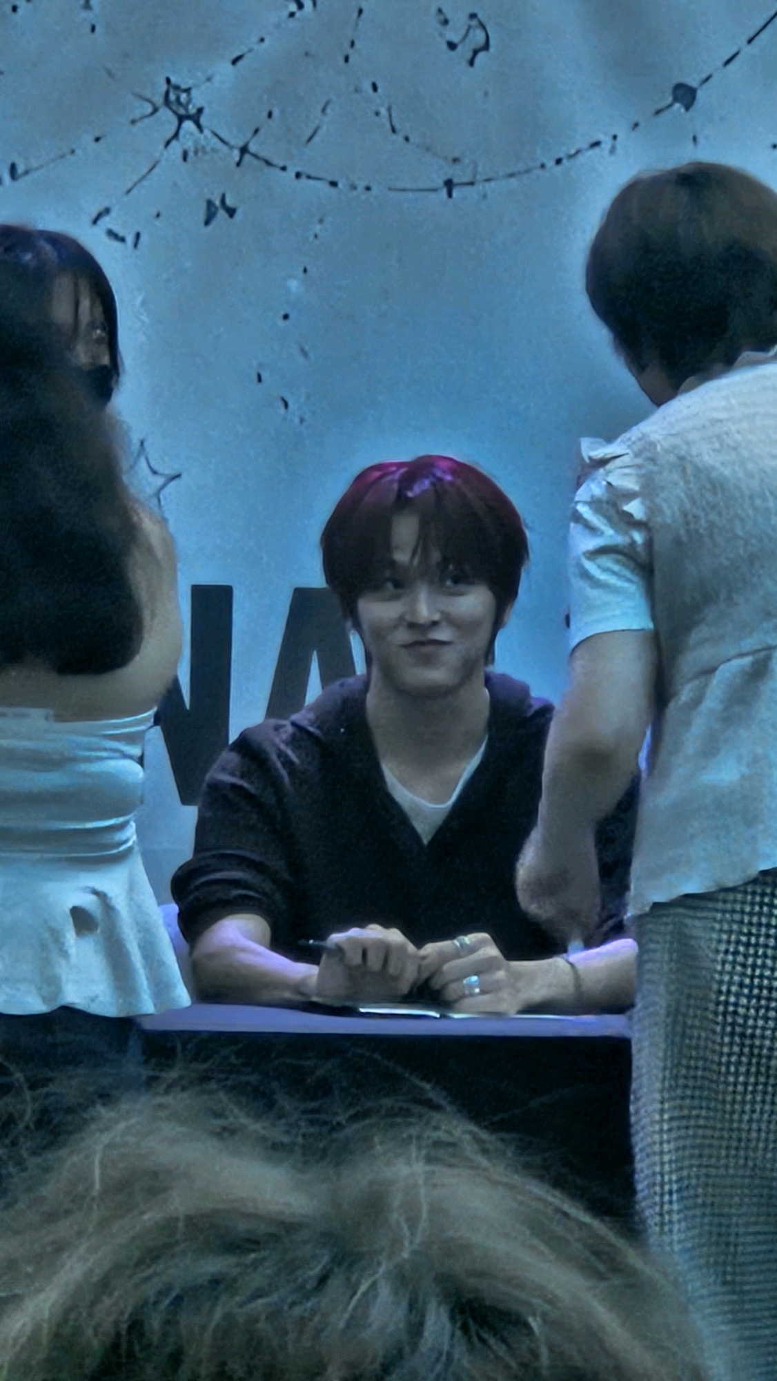 The image shows a man with short, dark red hair wearing a dark top over a white undershirt. He is seated at a dark table and appears to be signing something with a pen in his right hand. He is looking towards his right with a slight smile. Two individuals are partially visible on either side of him. On his left, a person with dark hair wearing a light-colored top is seen from the back. On his right, another person is visible from the back, wearing a patterned skirt and a light-colored top. The background is a light blue wall with some abstract dark markings. The foreground is blurred and appears to be the heads of a crowd. The scene suggests an autograph signing event. There is no visual information that places this scene in Barcelona, Spain.