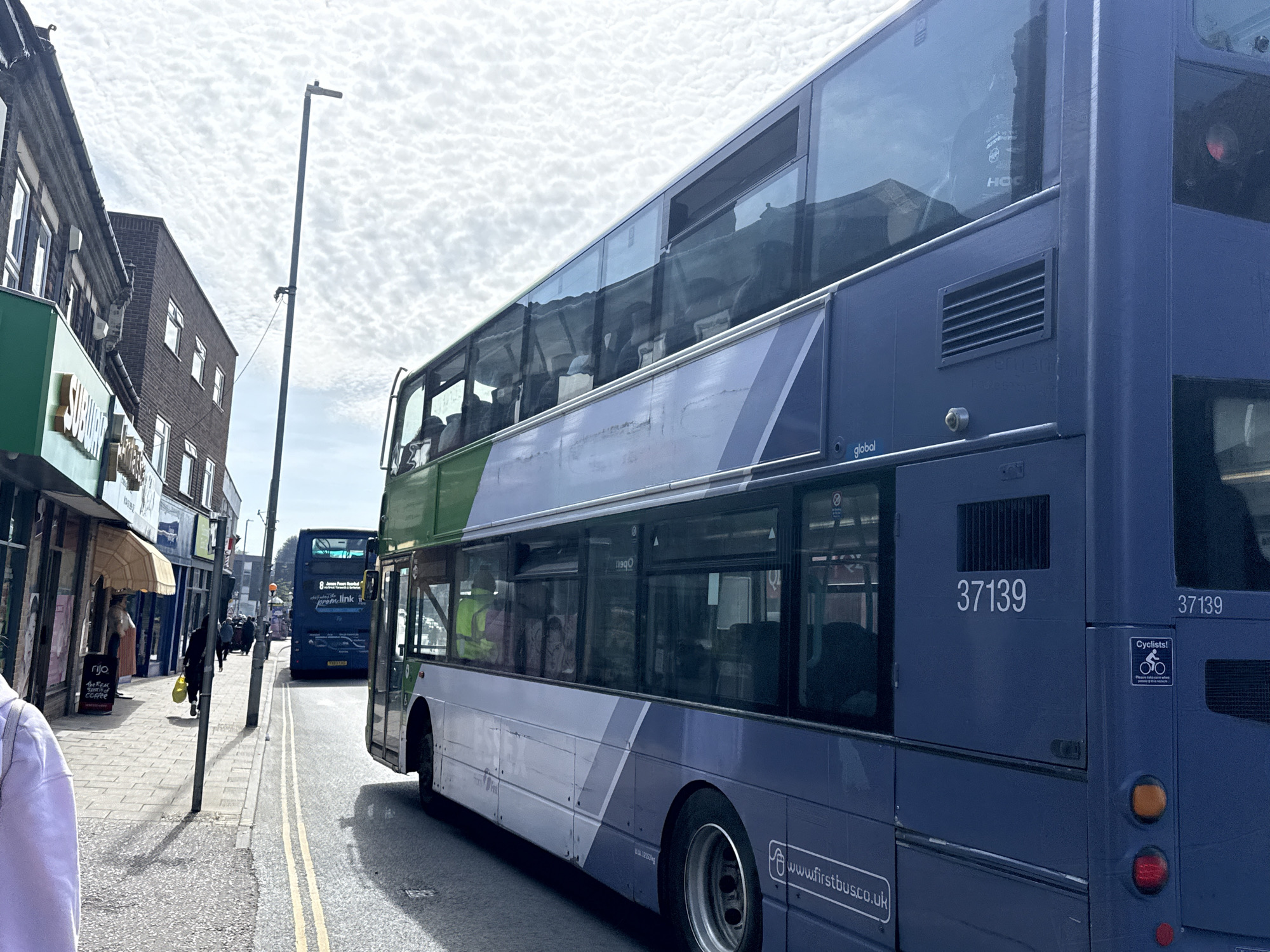 The image captures a street scene in Gorleston-on-Sea, United Kingdom, focusing on two First Bus double-decker buses. The bus in the foreground is a blue and white livery with green accents on the top portion of the body near the front. The number "37139" is prominently displayed on the rear of the bus, along with a warning sticker for cyclists. The website address "www.firstbus.co.uk" is also visible on the lower portion of the bus's side. Passengers can be seen inside the bus. Behind the first bus, another blue double-decker bus is visible, with the number "8" on the rear. People are walking on the sidewalk in front of shops and business buildings. Signage for "rijo" and "THE REAC coffee" can be seen. There is a street light next to the building with a "Subway" sign. The sky is overcast, with white cloudy light above. A double yellow line is marked on the street.