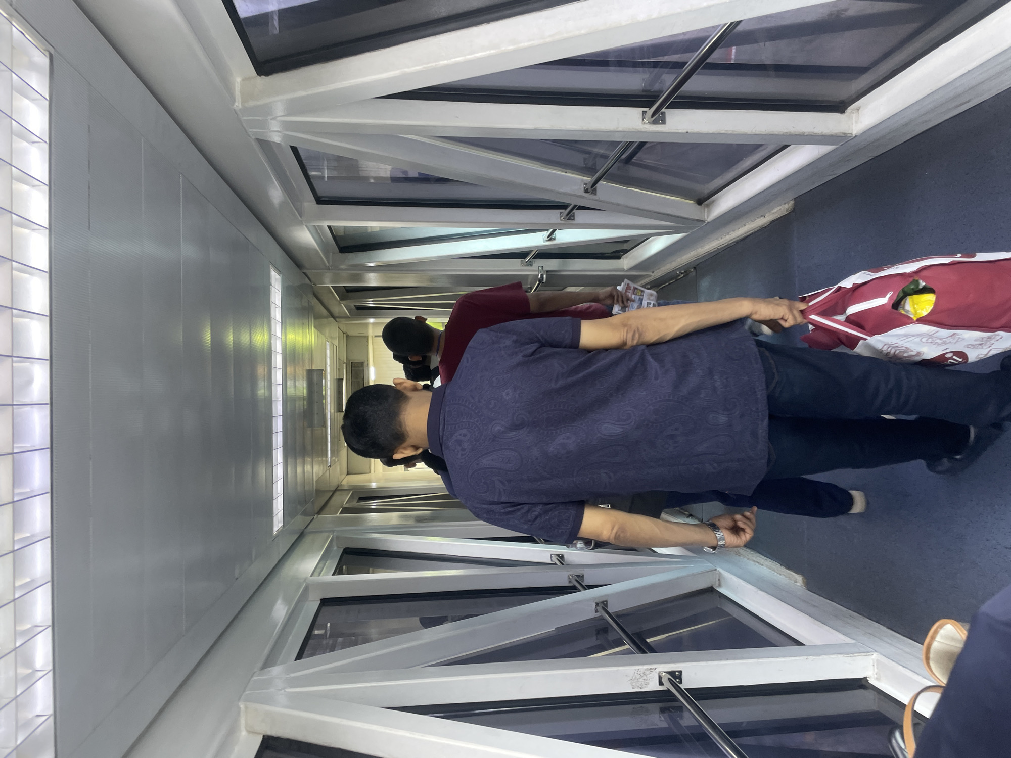The image depicts a group of people walking through a jet bridge, likely at Sardar Vallabhbhai Patel International Airport in Ahmedabad, India. The jet bridge has a white metal frame and large glass panels on both sides, offering a view of the exterior which is not clearly visible due to reflections and the angle. The ceiling is illuminated by fluorescent lights arranged in a grid pattern.
In the foreground, a man wearing a dark blue short-sleeved shirt with a paisley pattern is walking away from the camera. He has a watch on his left wrist and is carrying a red and white reusable shopping bag in his right hand, which appears to contain some yellow items. Behind him, another man in a dark shirt is visible, and further ahead, a person in a red t-shirt is walking. The floor of the jet bridge is a dark blue carpet. The overall scene suggests passengers boarding or disembarking an aircraft.