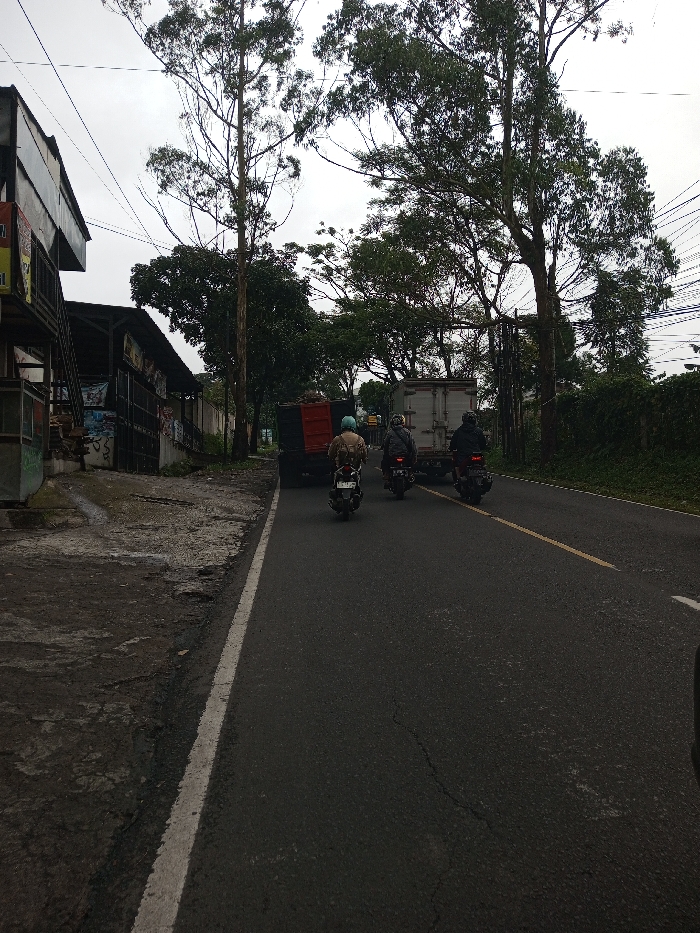 The image depicts a road scene in Serpang Shalli, Bhutan, under an overcast sky. The paved asphalt road features a faded yellow dashed center line and a solid white line marking its left edge. Three individuals are observed operating scooters or motorcycles, traveling in line away from the viewer within the right lane.
The lead motorcyclist, closest to the foreground, wears a light blue helmet and a tan backpack, riding a light-colored scooter. Immediately behind and slightly to the right is a second motorcyclist, wearing a dark helmet and dark clothing, operating a dark-colored scooter. The third motorcyclist, positioned furthest back in the line, also wears a dark helmet and dark attire, riding a dark scooter with an item secured to the rear.
Further down the road, two trucks are visible. The first is a black truck with a red cargo bed, positioned ahead of the motorcycles. The second is a white box truck, located further down the road and to the right of the black truck. Both trucks appear to be oriented in the same direction of travel as the motorcycles.
To the left of the road, an uneven, cracked concrete shoulder abuts a series of structures. The closest is a multi-level building with an open upper section displaying a red and yellow sign. Adjacent structures include a lower building with dark facade elements and posters, next to a concrete wall. The ground in this area consists of dirt and debris.
The right side of the road is bordered by a dark green, dense, vine-covered fence or wall. Numerous tall trees with varying foliage density line both sides of the road and extend into the background. Overhead, multiple power or communication lines crisscross against the gray sky, partially obscured by tree branches.