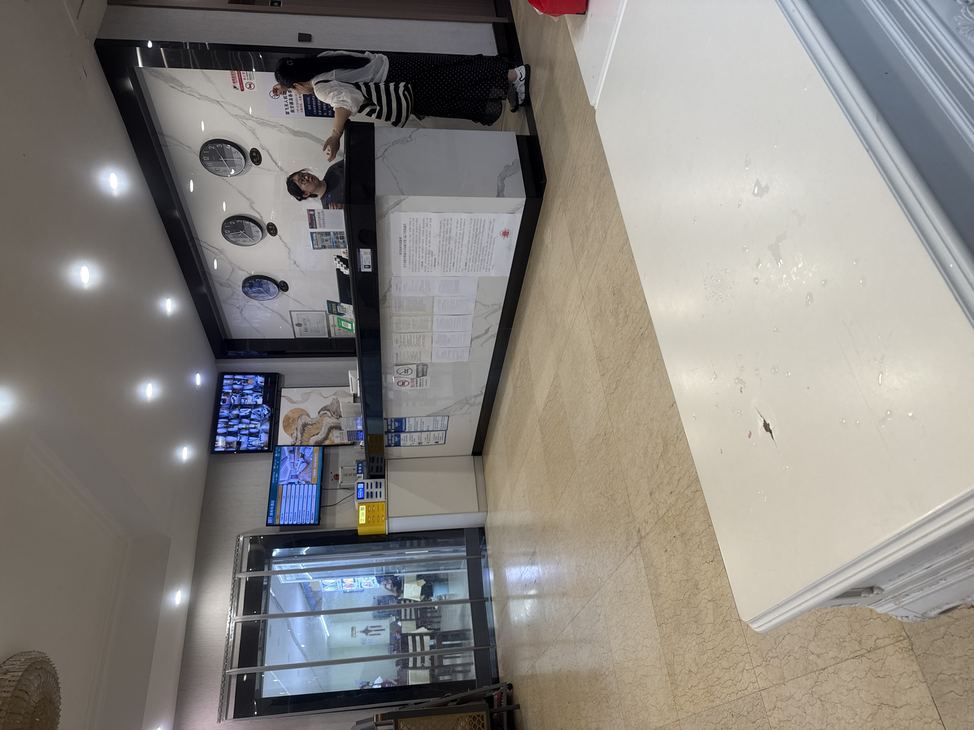 The image depicts the interior of a hotel lobby in Qinhuangdao, China. In the background, a reception desk is staffed by two women. One woman, wearing glasses and a grey t-shirt, appears to be speaking with a customer. The customer, a woman in a white top and black polka-dot skirt, gestures with her hands. Behind them, a marble-patterned wall features three large clocks displaying different time zones. Above the reception desk, multiple screens display security camera feeds and information relevant to hotel operations. To the left of the reception area, a set of glass doors leads to an adjoining space, possibly a dining area, with tables and chairs visible. The floor is tiled with a light-colored marble pattern. In the foreground, a white table with a slightly damaged surface is positioned centrally. The overall lighting in the lobby is bright, with recessed ceiling lights illuminating the space.