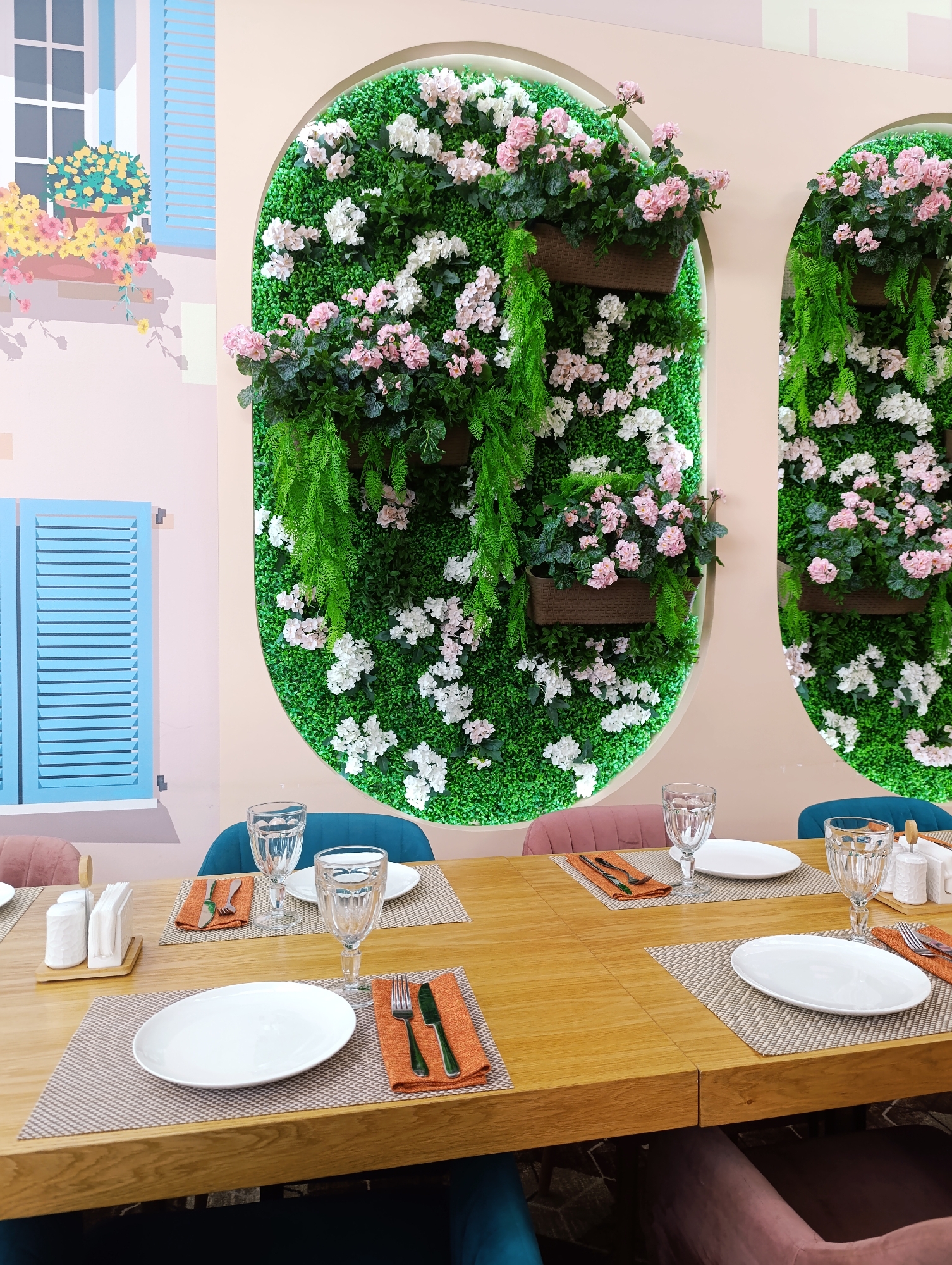 The image depicts a table setting in what appears to be a restaurant, possibly located in Unknown, Unknown. The table is wooden, set with woven placemats, white plates, silverware wrapped in orange napkins, and water glasses. Some of the settings also include salt and pepper shakers and napkin holders. The chairs surrounding the table are upholstered in various colors, including teal and mauve. Behind the table, the wall is painted with a pastel color scheme and features decorative elements, including a mural of a window with flowers and arched green walls with white and pink flowers. There are no people visible in the image, only hints of individuals seated at the table.