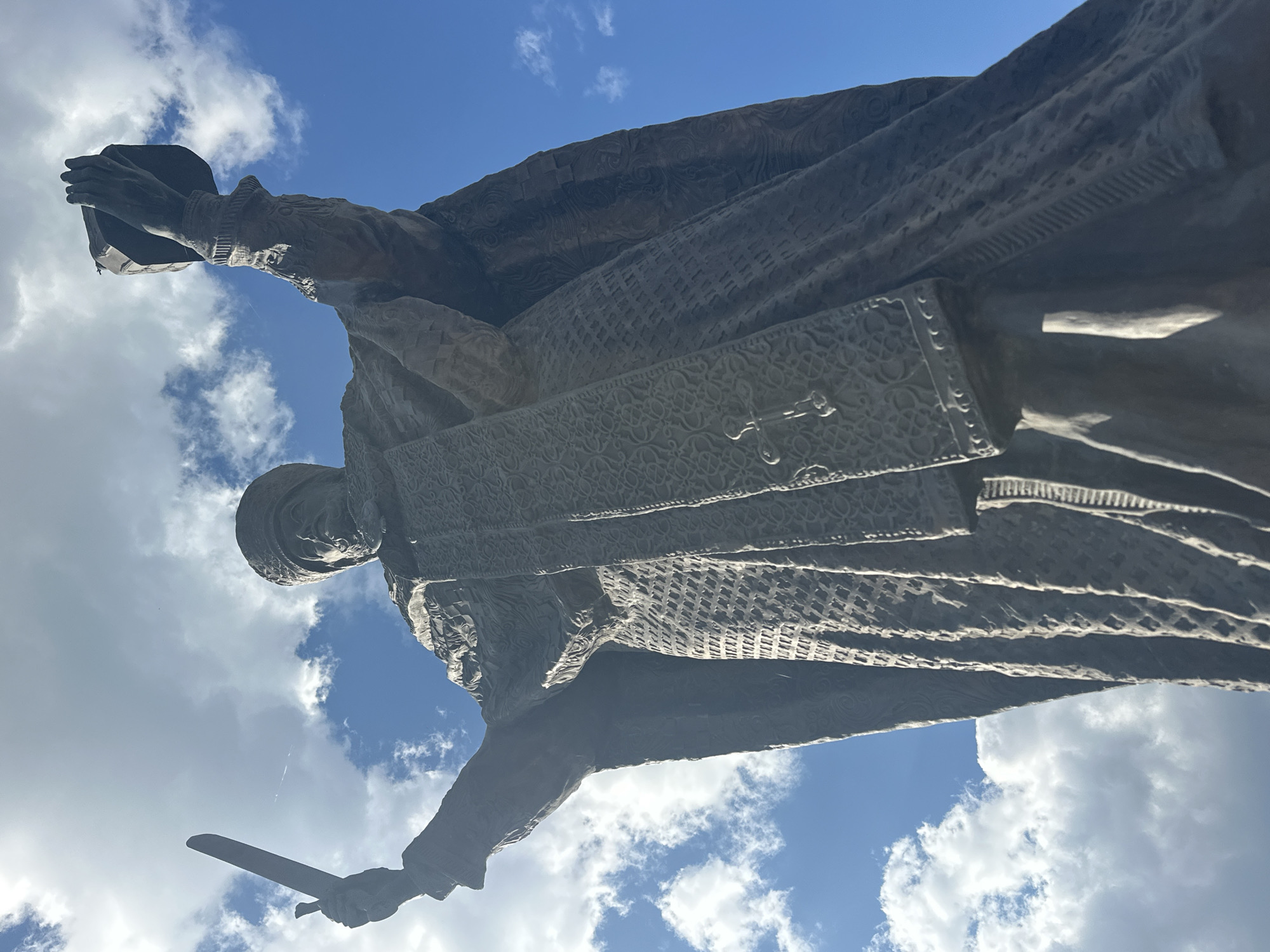 The image depicts a low-angle view of a statue against a backdrop of blue sky with scattered white clouds. The statue is of a male figure in historical clothing, including a hat, a long patterned robe, and what appears to be a decorative shield or panel on the chest. In one hand, the figure holds what looks like a book or tablet, and in the other hand, a sword. The statue is presumably located in Novosibirsk, Russia, based on the prompt's instruction. The perspective suggests the photograph was taken from the ground, looking up at the statue.