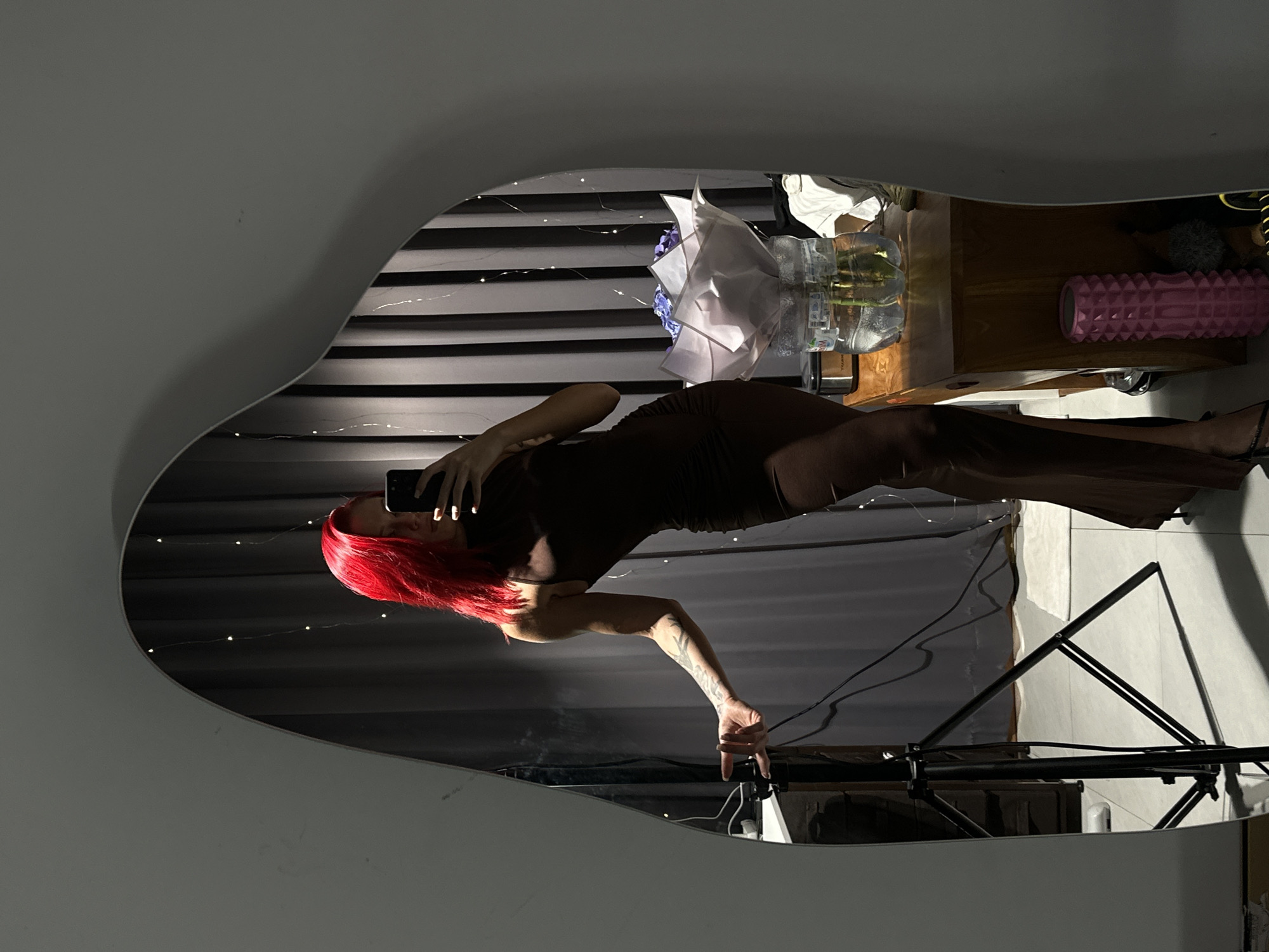 A person with red hair stands in front of an irregularly shaped mirror, holding a cell phone to take a selfie. The person wears a dark outfit with a form-fitting top and wide-leg pants. The reflection in the mirror shows a room with horizontal blinds, decorative string lights, and a bedside table with various items on top, including flowers in a vase and a pink foam roller. A tripod stands in the corner. The location is inferred as a personal indoor space. The city of Dalung, Indonesia, cannot be verified from this image alone.