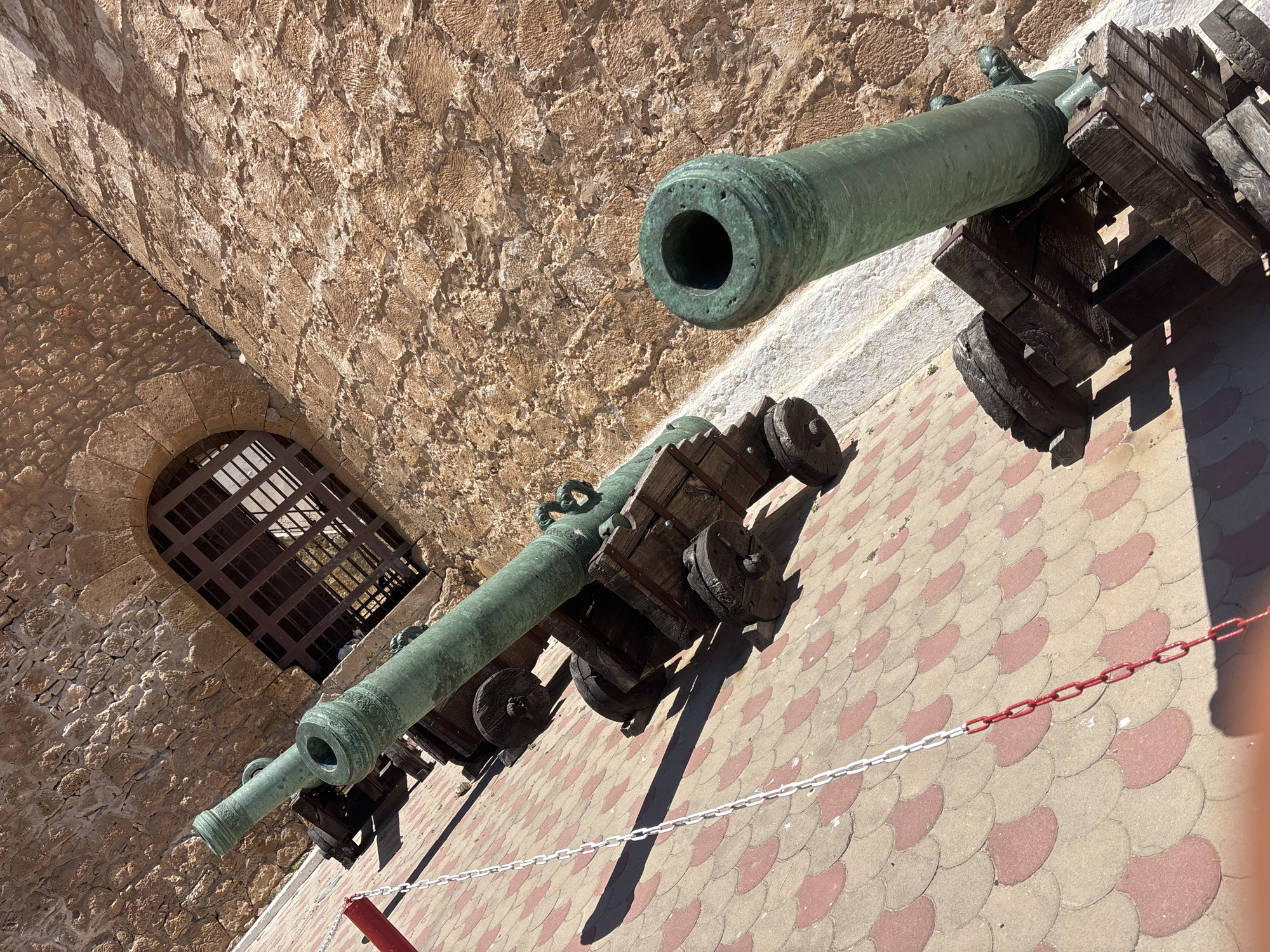 This image shows a row of antique cannons on their wooden carriages, positioned along a stone wall in a city square. The cannons are made of a greenish-bronze material and appear to be very old. They are arranged diagonally from the foreground to the background, with the closest cannon in the lower right corner. The wall behind the cannons is made of rough, textured stone blocks and has a large, arched opening covered by a metal grate. The ground in front of the cannons is paved with interlocking red and tan hexagonal tiles. A chain, partially painted red and white, runs along the paving stones in the foreground. The scene is brightly lit by sunlight, casting shadows on the ground and wall. The overall impression is of a historical site, possibly a fortress or defensive structure.