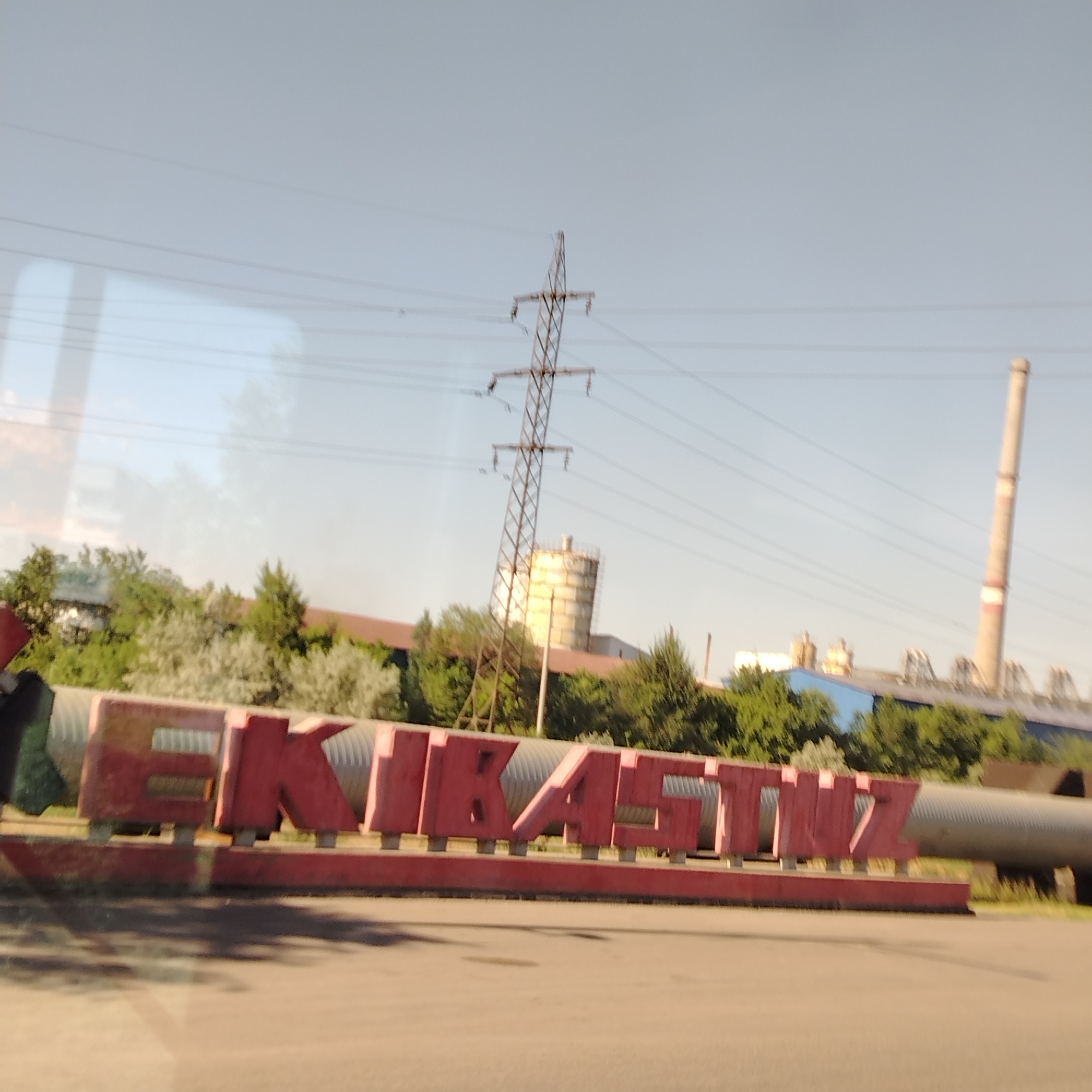 The image captures an industrial landscape under a clear sky. In the foreground, large, red, block-style letters spell out "EKIBASTUZ," serving as a prominent sign. Behind the sign, a large, corrugated pipe runs horizontally, and further back, a dense line of green trees and lighter foliage forms a natural barrier. Beyond the trees, industrial structures are visible, including a tall, slender smokestack with red bands and a cylindrical industrial building. A large electricity pylon with multiple wires crisscrossing the sky is also a significant element in the mid-ground. The overall scene suggests an industrial area within the city of Ekibastuz, Kazakhstan, given the prominent signage. The lighting indicates it is daytime, with sunlight casting shadows on the ground. The image appears to be taken from a moving vehicle due to the slight blur and the angle of the shot.