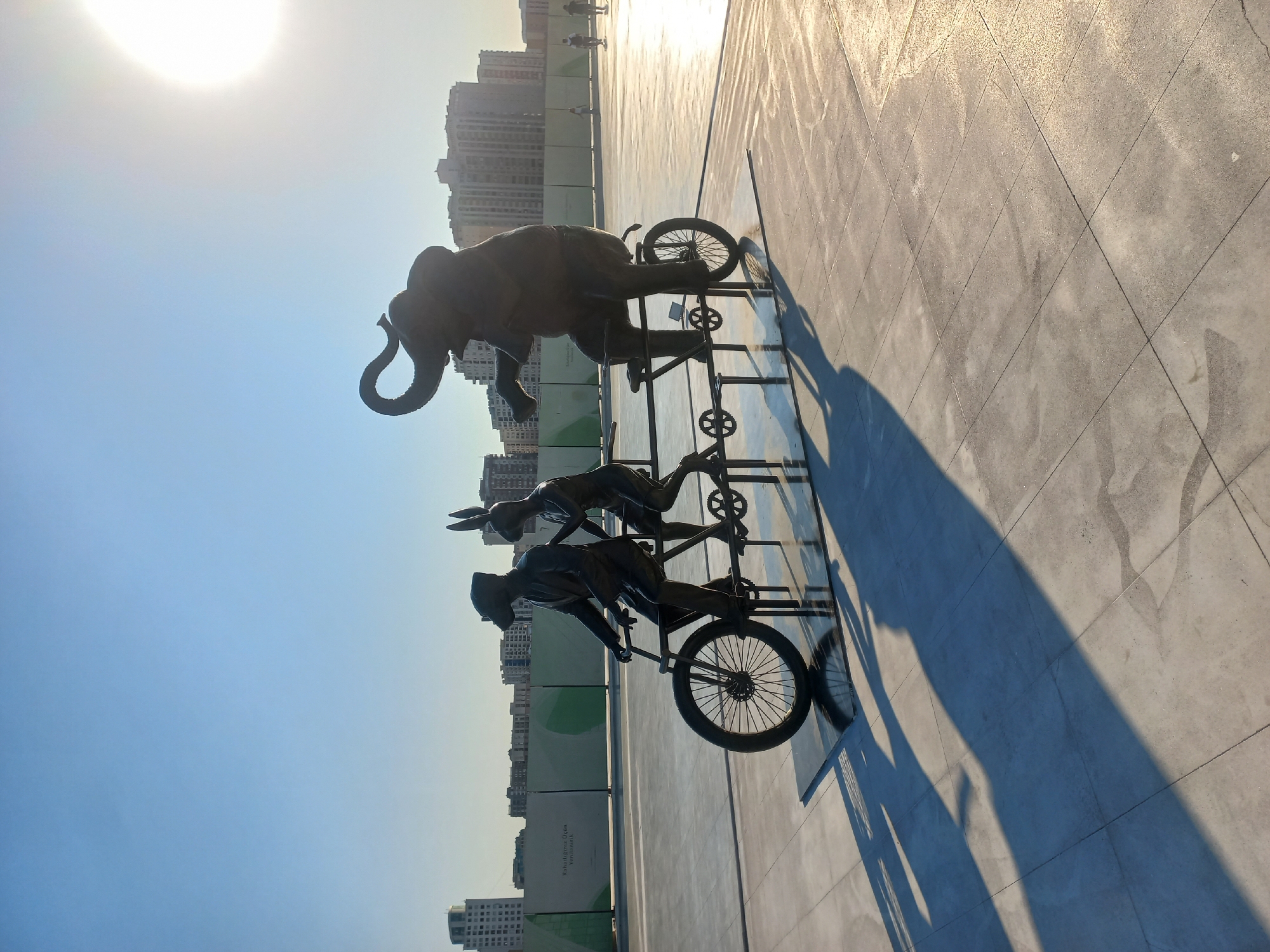 A bronze sculpture of three anthropomorphic animals on a tandem bicycle is situated on a paved outdoor plaza in Baku, Azerbaijan. The sculpture depicts a dog-like figure in the front, a rabbit-like figure in the middle, and an elephant in the back, all appearing to pedal the bicycle. They are positioned on a large, light-colored tiled surface with a reflective black base underneath the sculpture. The background features a low green fence with some text, followed by tall, modern buildings characteristic of a city skyline. The sky is clear and bright blue, with the sun positioned in the upper right corner, casting long shadows from the sculpture across the plaza. The scene is illuminated by the warm light of a setting or rising sun, suggesting it is either early morning or late afternoon. There are a few blurry figures in the distance, walking or skating on the plaza. This location is identifiable as the Baku Boulevard, a popular waterfront promenade.