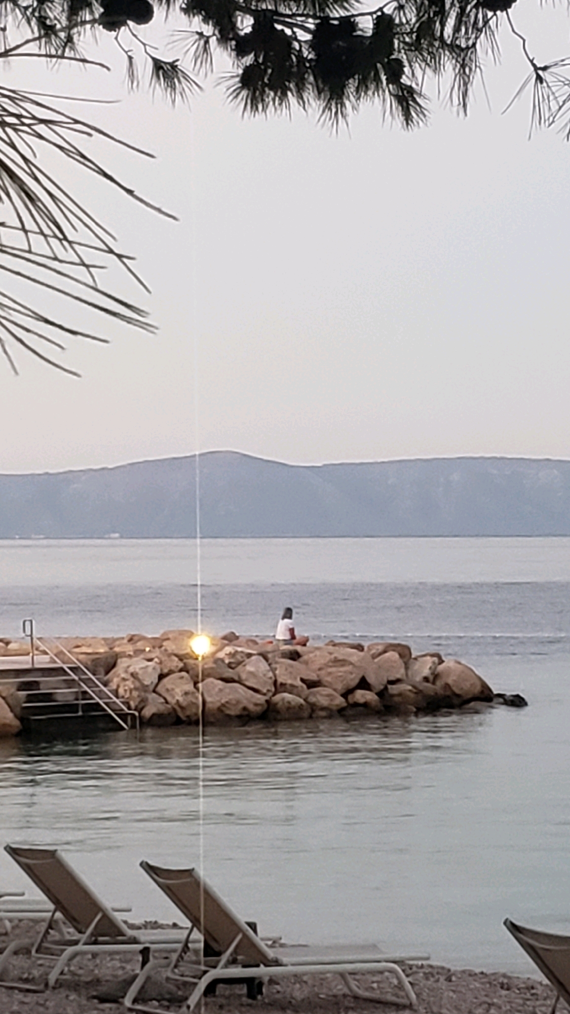 An outdoor scene in Općina Tučepi, Croatia, depicts a tranquil coastal view. In the foreground, two empty lounge chairs are positioned on a pebble beach, facing the calm sea. The water reflects the overcast sky and the silhouettes of distant mountains. In the middle ground, a person sits on a rocky jetty extending into the water. The person is wearing a white top and red bottoms, and appears to be looking out at the sea. A bright light emanates from a lamp post on the jetty, casting a strong glow. Steps lead down from the jetty into the water. Overhead, dark pine branches with cones frame the top of the image. The overall atmosphere is serene and peaceful.