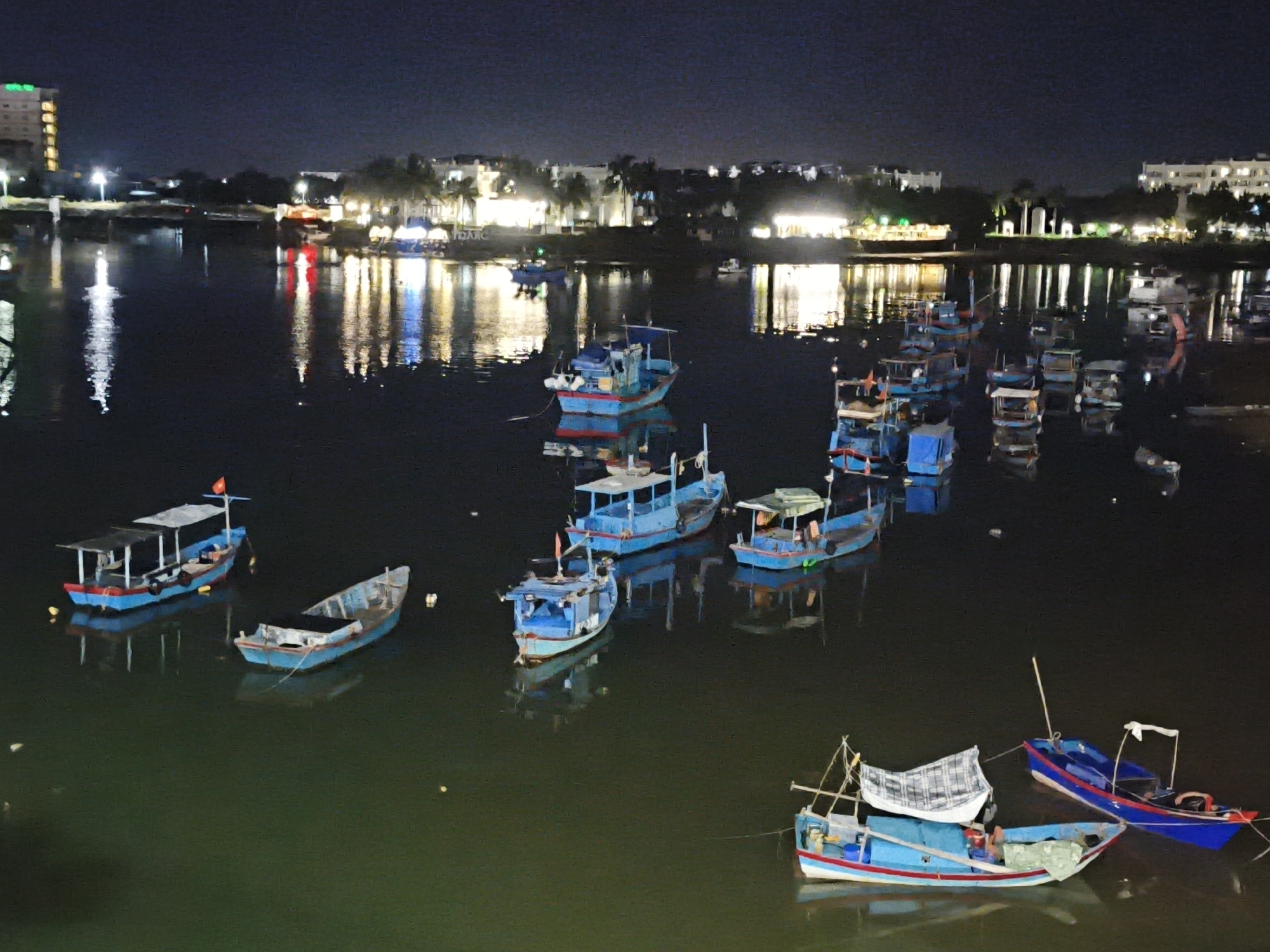 The image depicts a nighttime scene in the city of Bac Nha Trang Ward, Vietnam, showing numerous small fishing boats docked in a harbor. The water reflects the lights from buildings and streetlamps lining the shore, creating a vibrant, illuminated effect. The boats are a variety of colors, predominantly blue, with some featuring red accents and white canopies. Some boats appear to be loaded with equipment or supplies, while others are empty. The background shows a low-rise cityscape with illuminated buildings, suggesting a commercial or residential area. The overall atmosphere is calm and serene, with the gentle lapping of water and the distant glow of the city.