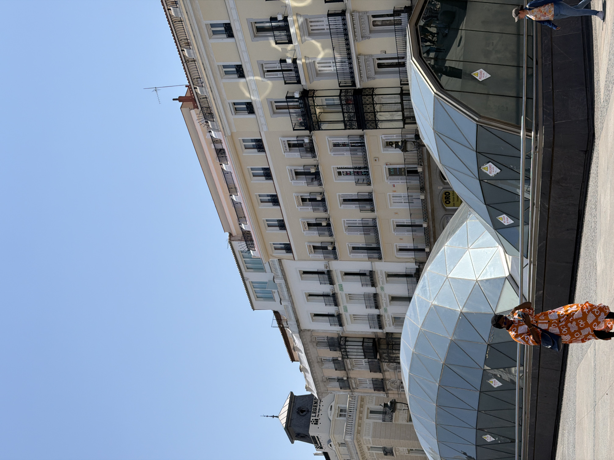 The image depicts a sunny day in Madrid, Spain, with a clear blue sky above. In the foreground, a modern, geodesic dome-like structure with glass panels is visible. A person in a vibrant orange and white patterned dress and dark pants, carrying a blue bag, walks towards the left, looking at their phone. Another person, wearing a cap, blue jacket, and jeans, walks towards the right. In the background, a row of historic, multi-story buildings with balconies, shutters, and ornate architectural details lines the street. One building features a tower with a sign that reads "LEGEND." The scene appears to be a public square or pedestrian area, possibly an entrance to an underground station, given the contemporary glass structure.