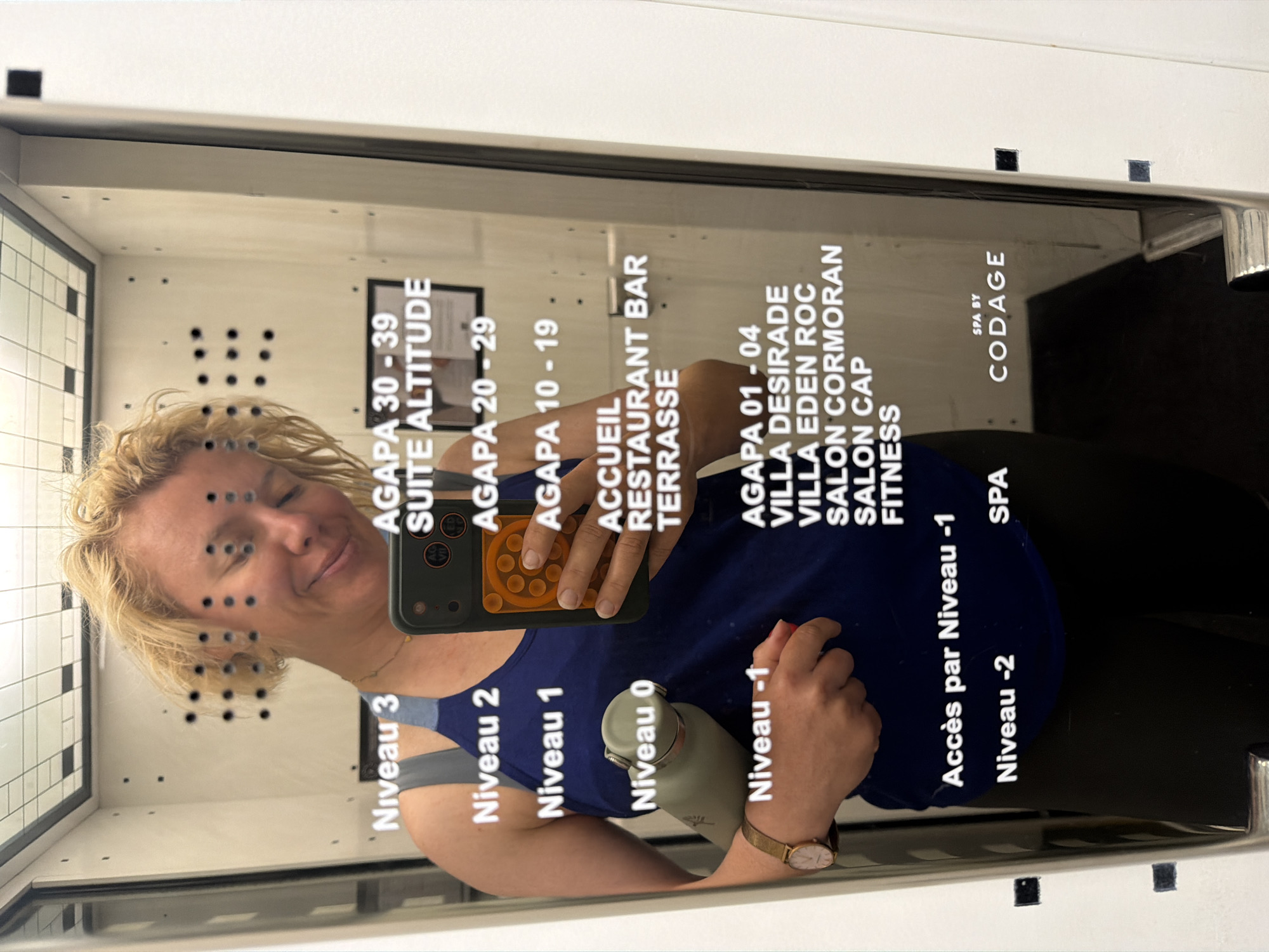 A blonde woman with her eyes closed and a smile on her face is taking a mirror selfie in what appears to be an elevator or a dressing room. She is wearing a blue tank top and black pants. She is holding a smartphone with a red pop socket and a green water bottle in her left arm. There are multiple lists of names and numbers on the mirror, including "Niveau 3", "AGAPA 30-39", "Niveau 2", "AGAPA 20-29", "Niveau 1", "AGAPA 10-19", "ACCUEIL", "RESTAURANT BAR", "TERRASSE", "Niveau -1", "AGAPA 01 - 04", "VILLA DESIRADE", "VILLA EDEN ROC", "SALON CORMORAN", "SALON CAP", "FITNESS", "Accès par Niveau -1", "Niveau -2", and "SPA BY CODAGE". The woman is wearing a watch on her left wrist. The background is the interior of the elevator or room, with a tiled ceiling and white walls.
