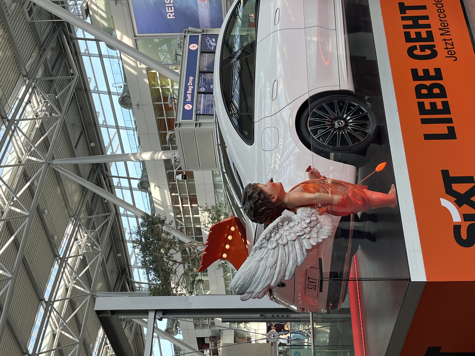 In an airport terminal in Dusseldorf, Germany, a pink Mercedes-Benz car is displayed on a raised platform. The car features a sculpture of a cherub with large white wings, holding a small orange arrow. The sculpture is positioned in front of the car, partially obscuring it. The car's license plate reads "SIXT". In the background, a "Self Bag Drop" sign is visible, along with an information board displaying flight details. The terminal has a modern architectural design with a metal roof structure and large windows. An orange banner at the base of the display reads "LIEBE GEHT Jetzt Mercedes".