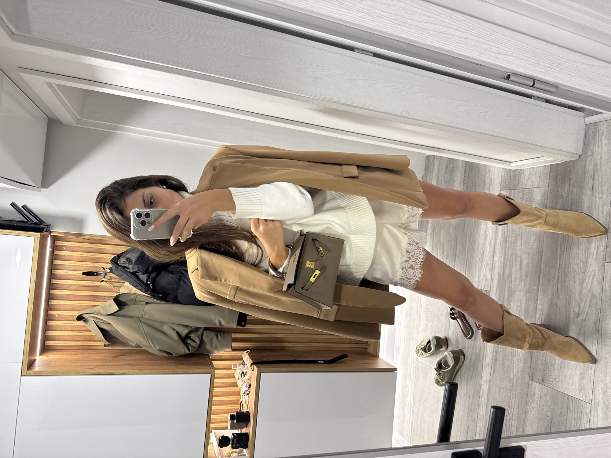 A woman is taking a mirror selfie in what appears to be a modern entryway or dressing area. She is wearing a white turtleneck sweater, white lace-trimmed shorts, a camel-colored blazer draped over her shoulders, and camel-colored suede cowboy boots. She is holding a small taupe handbag. In the background, a wooden slat wall with hangers holds a tan jacket and a black puffer jacket. On a white cabinet to the left are various bottles and accessories. The floor is a light gray wood-look tile. Scattered on the floor are a pair of green sneakers and a pair of brown slide sandals. The overall impression is one of curated style and personal presentation. There is no information in the image that allows for the identification of the city of Burgas, Bulgaria.
