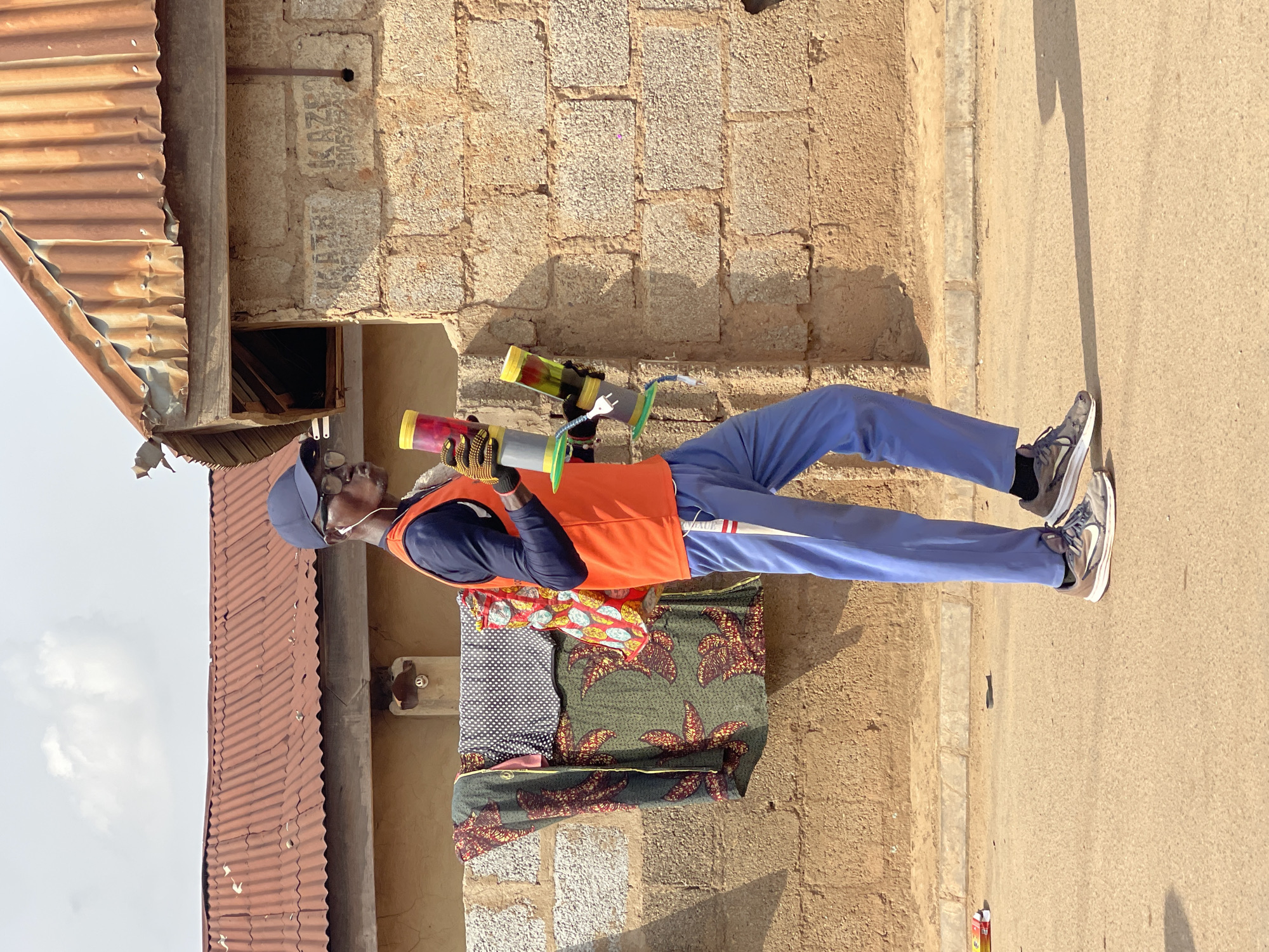 A man stands on a sunny day in Kuchingoro, Nigeria, appearing to be engaged in street vending. He is dressed in a blue cap, a dark blue long-sleeved shirt, a bright orange sleeveless vest, light blue trousers, and sneakers. He also wears dark gloves with yellow accents and glasses. In each hand, he holds a colorful, custom-made cylindrical device; the one in his left hand is red and yellow, while the one in his right is grey and yellow, featuring visible wires and an electrical plug. Beaded bracelets adorn his right wrist.
Behind the man, folded patterned fabrics, possibly merchandise, rest against a light brown, weathered stone wall. Faint text, resembling "KAZ" or "KAZO," is visible on some of the stone blocks. To the left, a section of a rusty corrugated metal roof is visible, suggesting a simple structure, with a dark opening beneath it. The ground to the man's right is a light tan asphalt or dirt road, bordered by a concrete curb. Strong shadows cast by the man and his objects indicate bright daylight. A small red and yellow box is on the ground near his feet.