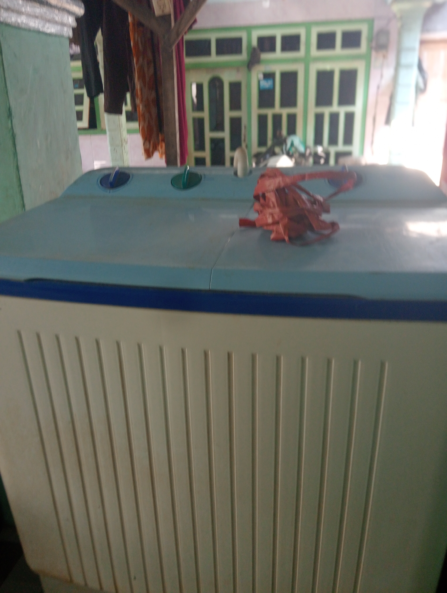 A top-down, medium shot captures a light blue twin tub washing machine in what appears to be an outdoor or semi-outdoor space in Besuki, Indonesia. The washing machine's lid is closed, and a bundle of red fabric is placed on its surface. Two circular knobs, one blue and one green, are visible on the top left of the lid. The front of the washing machine is a light cream color with vertical ribbed detailing. In the background, a building with green-framed windows and doors is visible. Laundry hangs on a wooden structure to the left of the washing machine. The overall scene is slightly blurred and has a soft, natural light.