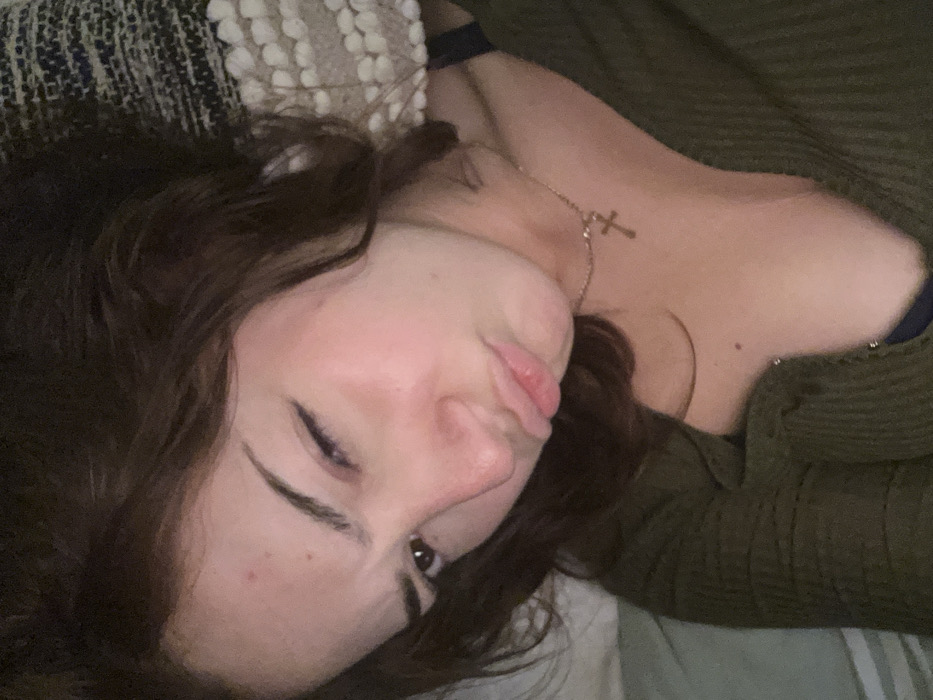 A woman with brown, wavy hair lies on her side, face close to the camera. Her lips are puckered. She wears a green, textured cardigan and a delicate necklace with a cross pendant. Part of a patterned pillow is visible behind her head. The lighting is soft, illuminating her face and neck. Her eyebrows are dark and arched. She is resting on what appears to be a bed with white sheets.