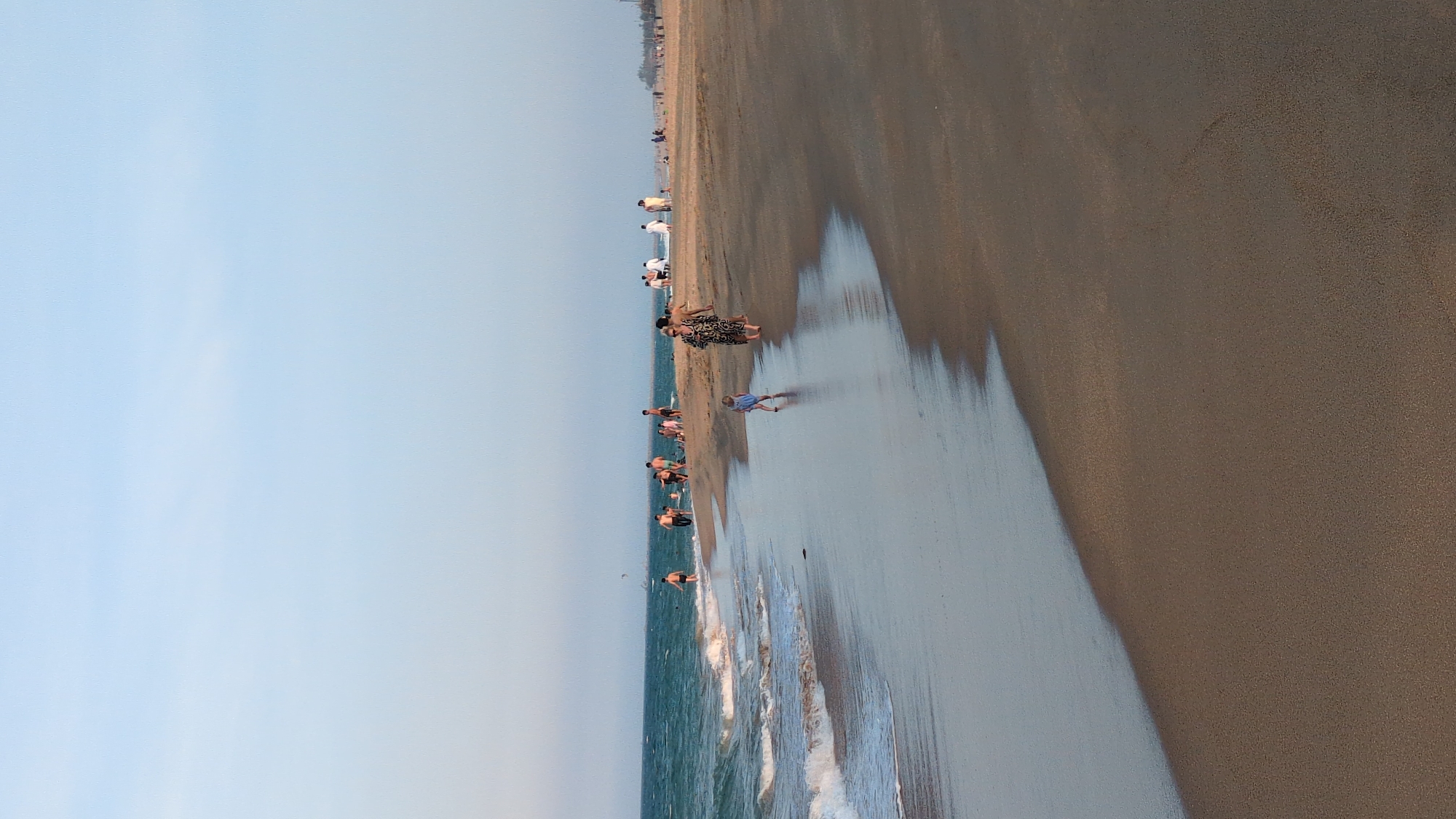 The image depicts a beach scene in Tuy Hòa, Vietnam. Along the shoreline, several people are visible: some are wading in the shallow water, while others are standing or walking on the sand. The beach has a broad sandy area next to the water and a lighter expanse reflecting a low-angled light. The sky is pale blue.