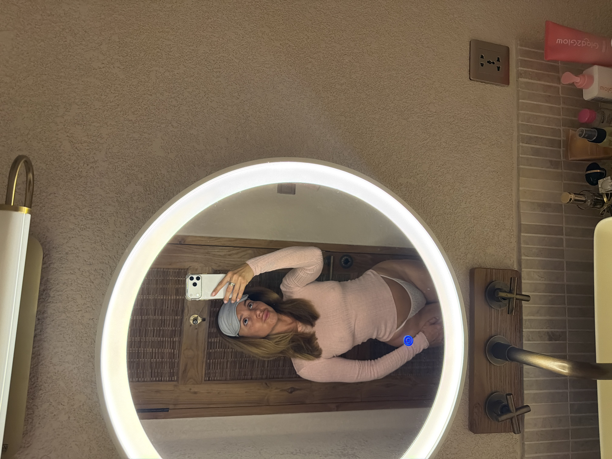 A light-skinned woman with blonde hair is reflected in a lighted round mirror in a bathroom. She is wearing a pink long-sleeve shirt, white underwear, and a gray sleep mask on her forehead. She holds a white cell phone up to the mirror with her right hand, presumably taking a selfie. The bathroom appears to be in a hotel or resort, with a wooden cabinet behind the mirror and tiled walls to the right. The location is likely in Mengwi, Indonesia, based on the environment. A power outlet is visible above the mirror, and various toiletry items are on a shelf to the right. A wooden panel with three brass knobs and a faucet is to the right of the mirror.