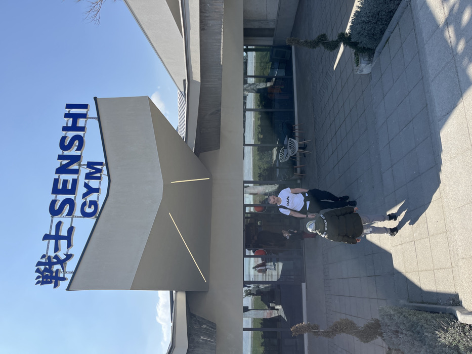 The image presents the exterior of the "SENSHI GYM" building in Varna, Bulgaria, under a clear, bright blue sky. The building exhibits modern, angular architecture with light-colored facades and extensive glass panels at ground level. A large blue sign on the roof displays "戦士 SENSHI GYM". Below the sign, a canopy projects over the entrance, featuring integrated linear light fixtures.
Two individuals are depicted walking away from the camera on a paved forecourt composed of light grey tiles. The lead person, a child or young adolescent, wears a dark olive puffer jacket with "MONCLER" visible on the left sleeve, light grey sweatpants, and dark shoes. Following this individual is an adult male, wearing a white t-shirt with "RAW." printed on the chest, black trousers, and a white garment tied around his waist. Both individuals cast distinct shadows on the pavement, indicative of direct sunlight.
The glass panels of the building's entrance reflect a green, possibly grassy area and a body of water beyond. To the right of the entrance, two white wicker chairs and a small round table are arranged as outdoor seating. Small, manicured shrubbery and a potted spiraled topiary plant are visible on the periphery of the paved area.
