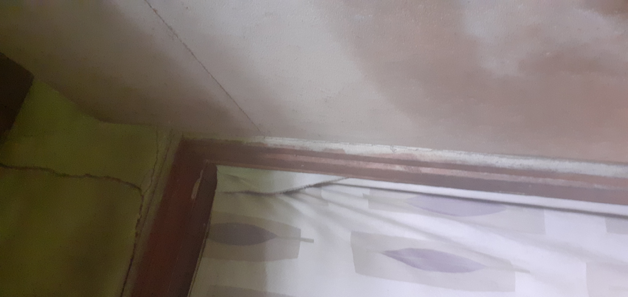 The image shows a corner of a room. The ceiling is a light beige color with a crack running across it. The wall on the left is light green with visible cracks. A dark brown wooden frame is visible at the corner, possibly of a window or a doorway. Inside the frame, a white curtain with light purple leaf-like patterns can be seen. The location is Alasia, Nigeria.