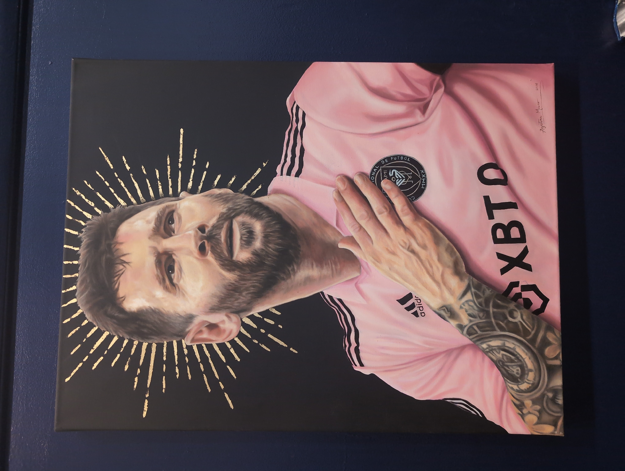 This is a portrait painting of Lionel Messi, a professional soccer player, hanging on a dark blue wall. The painting depicts Messi from the chest up, wearing a pink Inter Miami CF jersey with black stripes on the shoulders and Adidas branding on the chest. He has a beard and is looking off to the side with his right hand placed on his chest. Behind his head is a gold sunburst halo design. The painting is signed in the bottom right corner with "Agustina Mourao" and the year "2026". The painting is framed and mounted on the wall. The lighting suggests it's indoors.