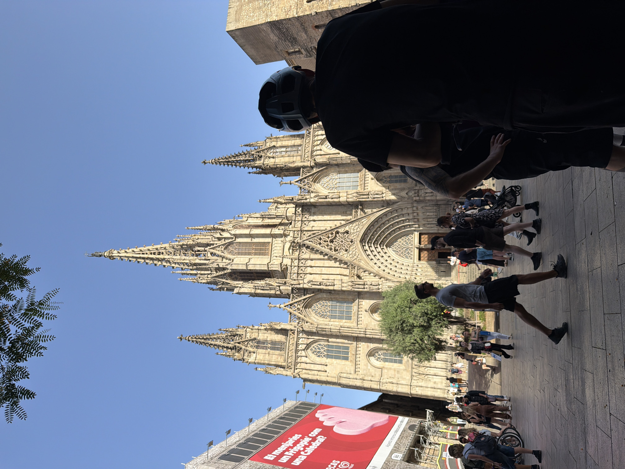 The image captures the facade of the Barcelona Cathedral, a prominent Gothic cathedral located in the heart of Barcelona, Spain. The cathedral's intricate spires and detailed stonework are bathed in sunlight, set against a clear blue sky. In the foreground, a bustling public square is visible, with several people walking and interacting. Some individuals are silhouetted against the bright background, including a person wearing a helmet, possibly a cyclist or pedestrian. A large billboard with red and white lettering is visible on the left side of the image, adding a contemporary element to the historical setting. The pavement is made of cobblestones, characteristic of many European city squares. The scene depicts a vibrant urban environment where historical architecture and modern life coexist.