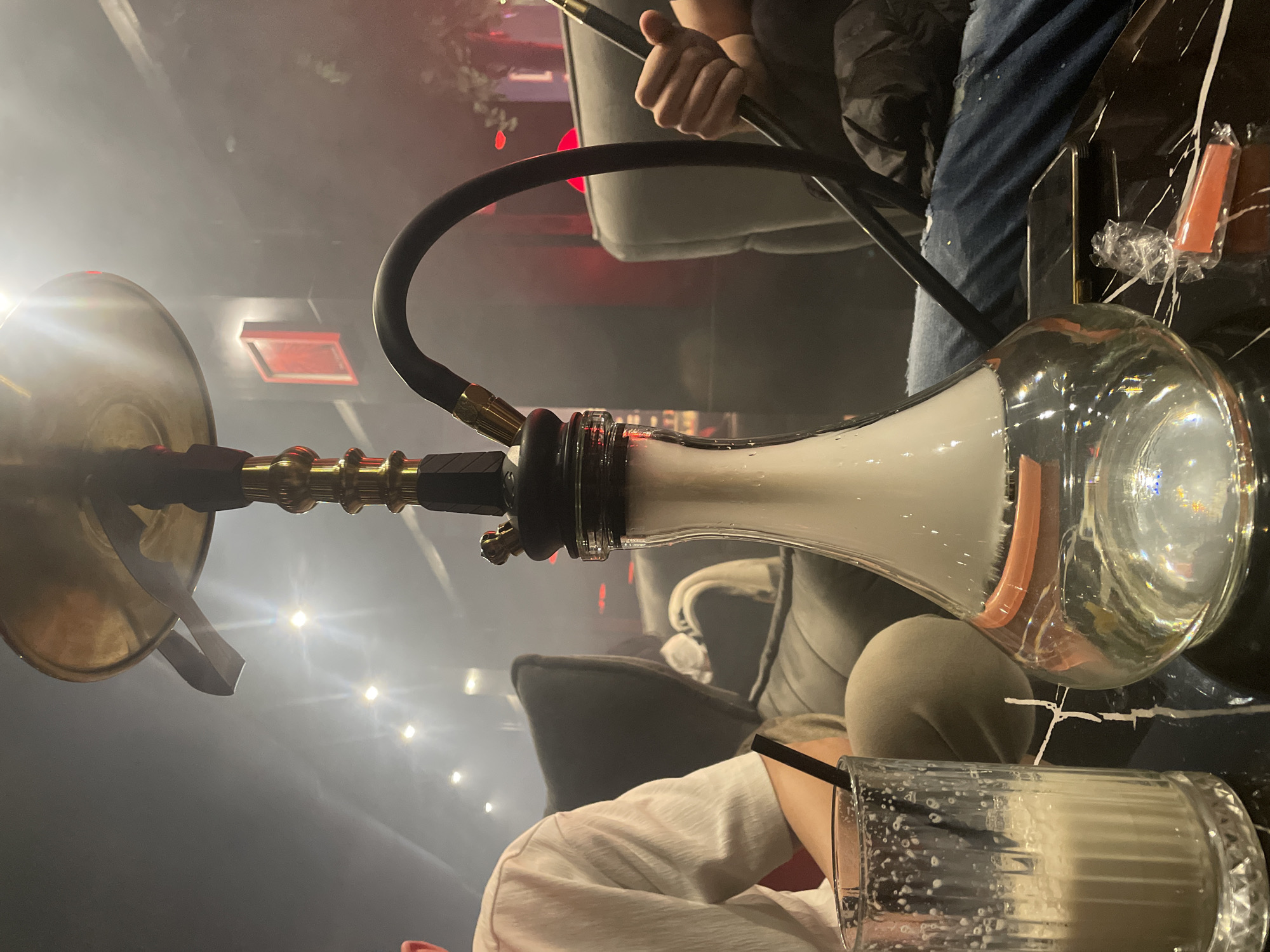 The image captures a hookah setup on a dark, reflective surface, likely a table. The hookah has a brass top and a glass base filled with white smoke. A black hose connects the top to the base. In the background, a person is holding the hookah's hose with their right hand. Another person is seen in the lower right corner, wearing a white shirt and holding a glass with a black straw inside. The setting appears to be indoors with soft lighting. The location is the city of Varna, Bulgaria.