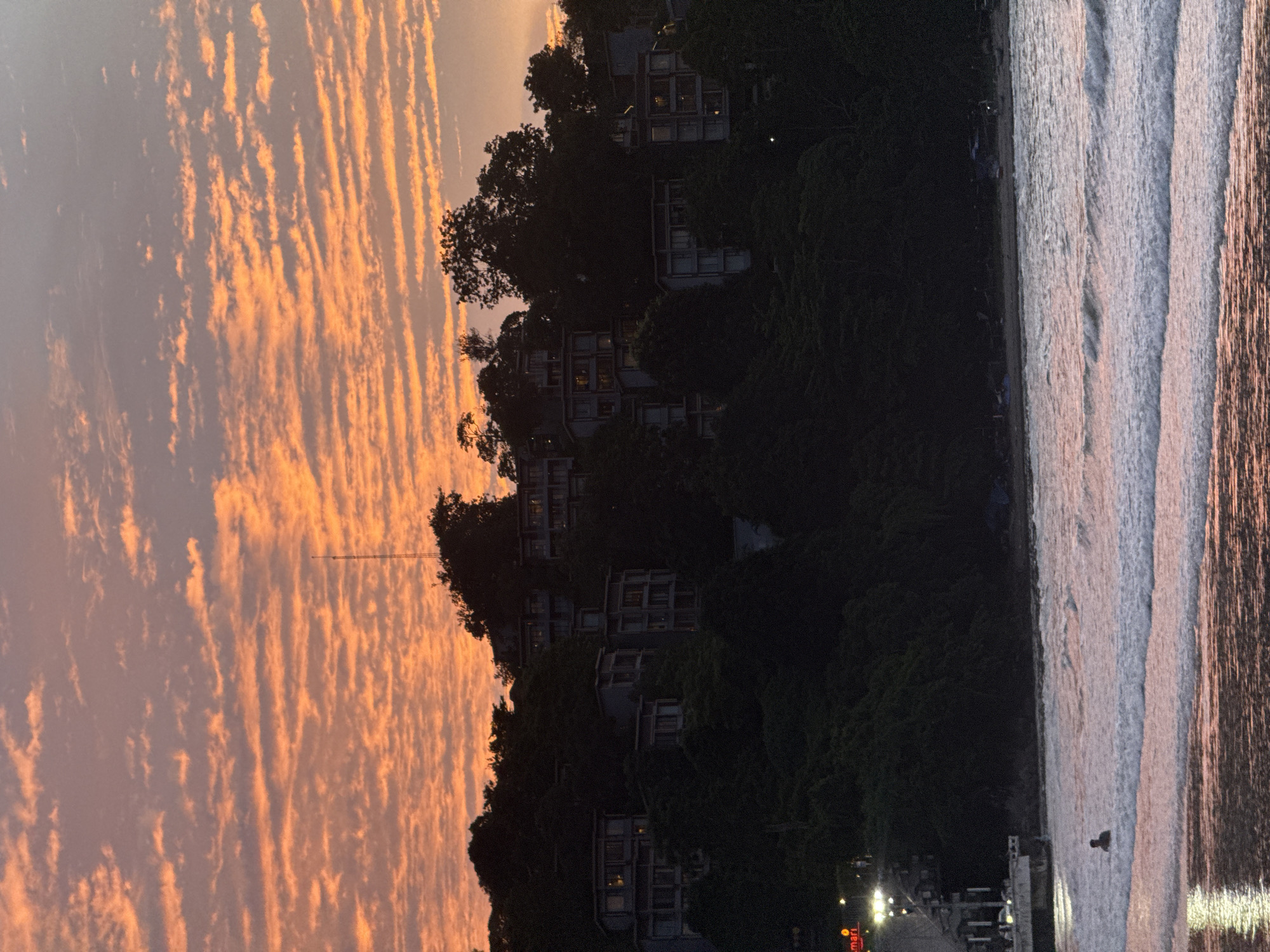 The image shows a coastal view of Patong, Thailand, during sunset. In the foreground, waves are breaking on a beach, and a lone individual is visible wading in the shallow water. In the midground, a dark, tree-covered hillside is dotted with several multi-story buildings, likely hotels or resorts, illuminated by interior lights. The buildings are nestled into the dense foliage, suggesting a hillside development. The background features a vibrant sunset sky with scattered clouds painted in shades of orange and purple, indicating the end of the day. A tall radio tower is faintly visible against the sky on the highest point of the hill. The overall scene conveys a tranquil evening atmosphere at a popular tourist destination.