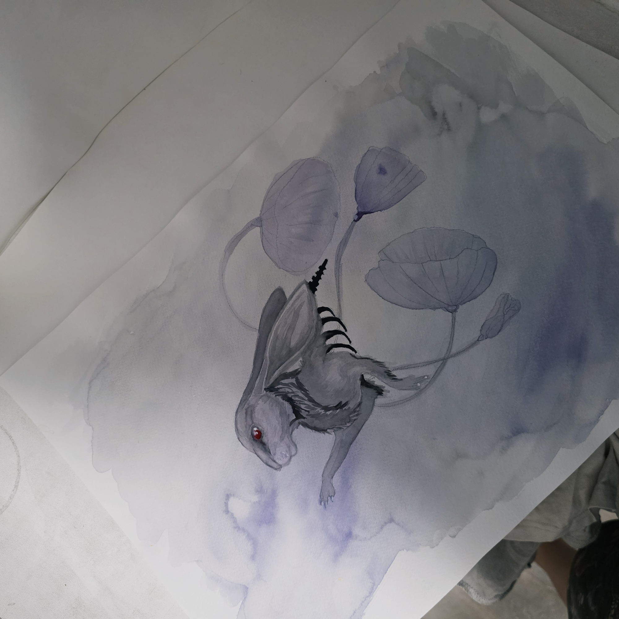 The image is a watercolor painting of a creature that appears to be a rabbit with a single red eye, striped back, and flowers around it. The creature is gray and positioned in the center of the image. The background is a mix of light and dark gray and blue watercolor washes. Three gray flowers, resembling poppies, with long, curved stems are arranged around the rabbit. The style is dreamlike and fantastical. The scene is indoors. The location is undetermined.